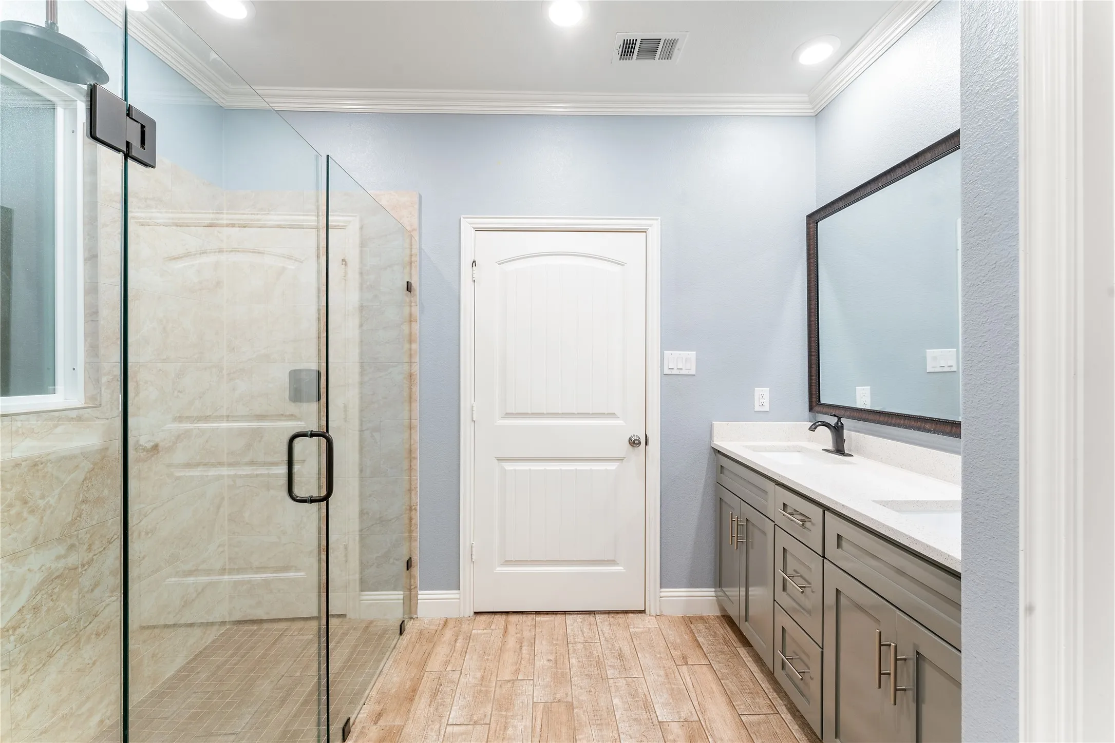 Spacious bathroom featuring a glass-enclosed shower, a double vanity with white countertops, light wood-finish flooring, and recessed lighting