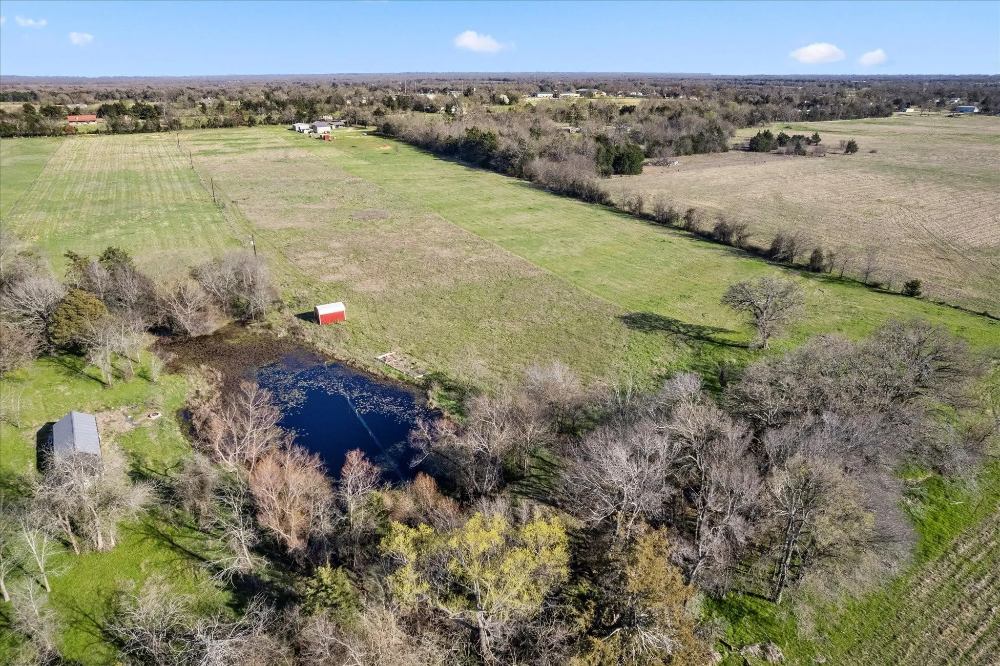 Expansive acreage featuring a natural pond, surrounding mature trees, a red storage shed, and a gray-roofed outbuilding