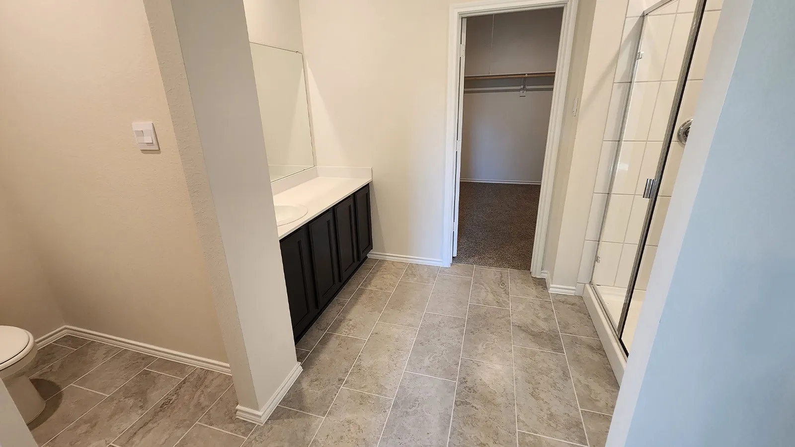 Bathroom featuring tile flooring, a dark wood-finish vanity with a white countertop and integrated sink, a frameless wall mirror, a commode, and a glass-enclosed shower