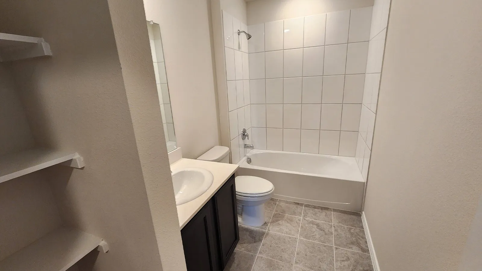 Full bathroom featuring a white porcelain tub and shower combination with white subway tile surround, a dark wood-finish vanity with an integrated sink, and gray tile flooring