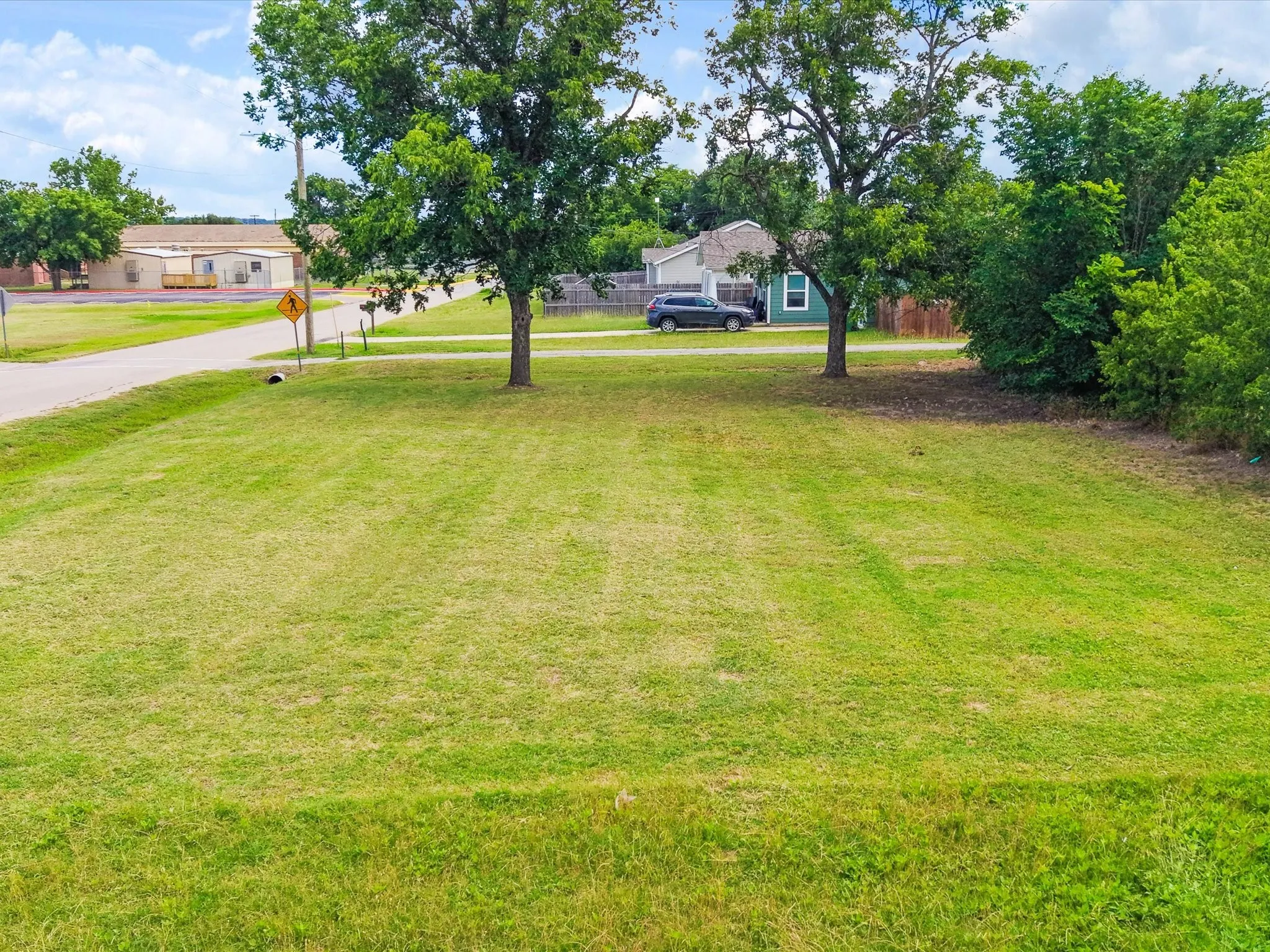 Expansive green lot featuring two mature trees and a paved road frontage