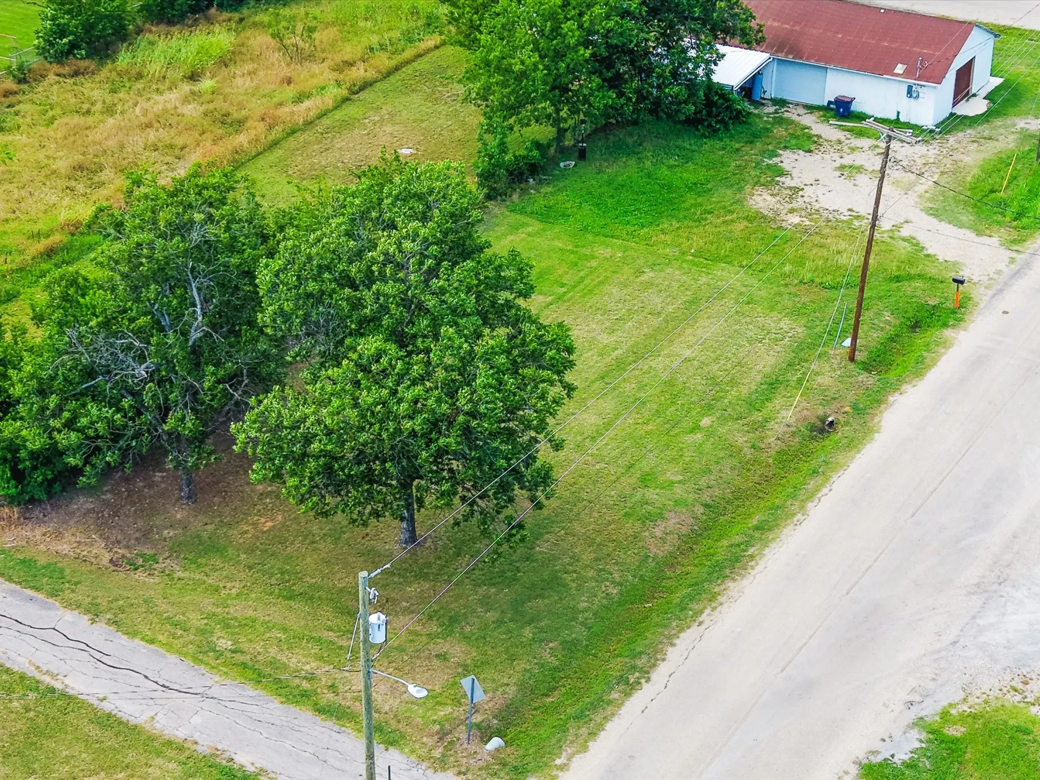 Expansive land parcel featuring mature trees, a partially paved road frontage, and a white commercial-style building with a red roof