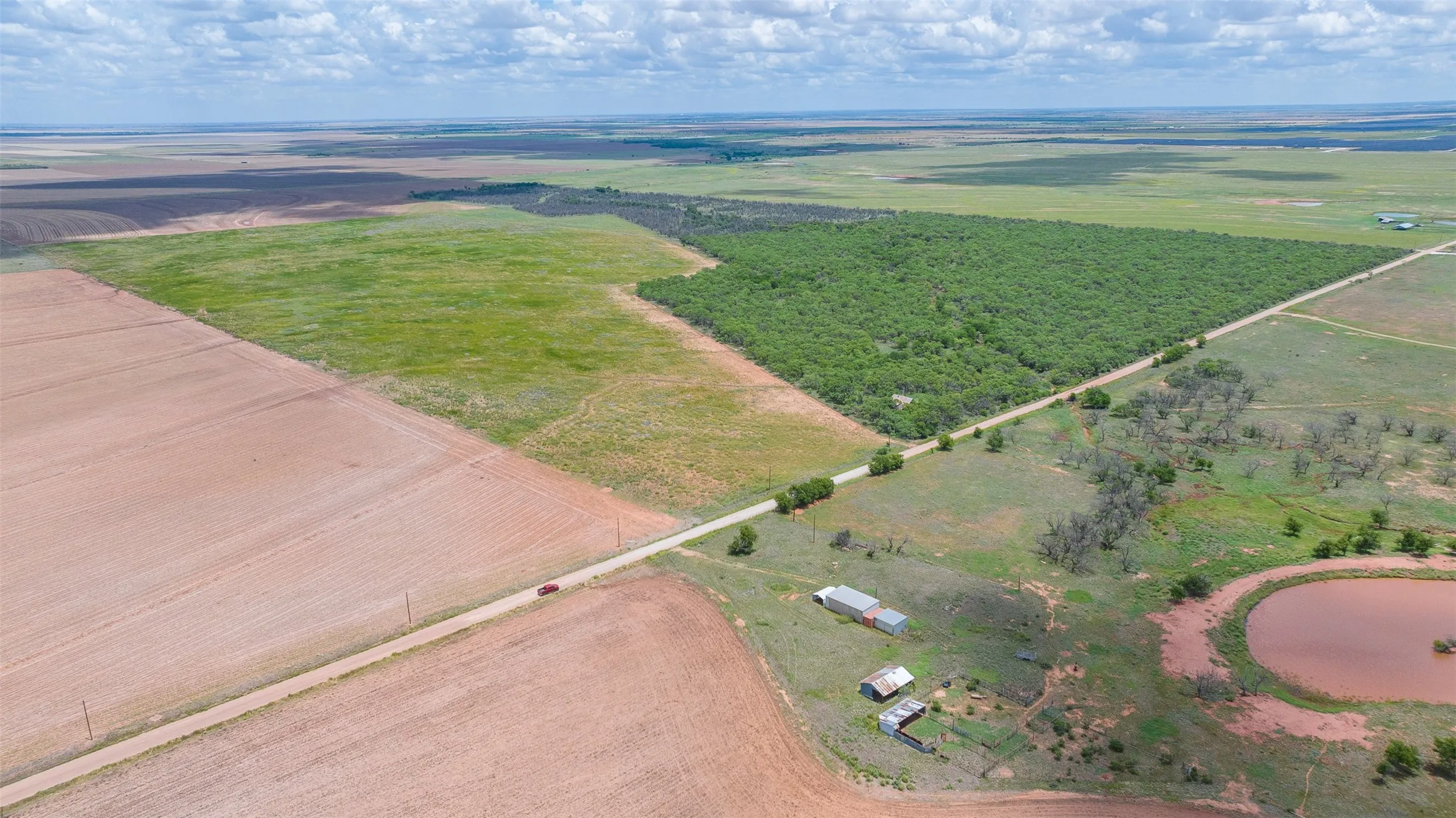 Expansive agricultural land featuring a verdant wooded area, cultivated fields, a dirt road traversing the property, a natural pond, and accessory structures