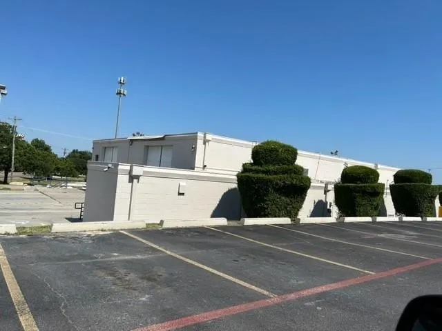 Commercial building featuring a light-colored exterior, an expansive paved parking area, and meticulously shaped topiary landscaping