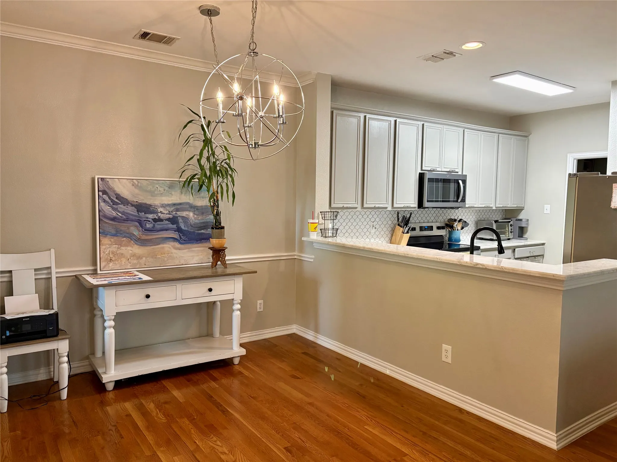 Hardwood flooring, crown molding, chair rail detailing, a contemporary chandelier, and a kitchen featuring white cabinetry, a tiled backsplash, and stainless steel appliances