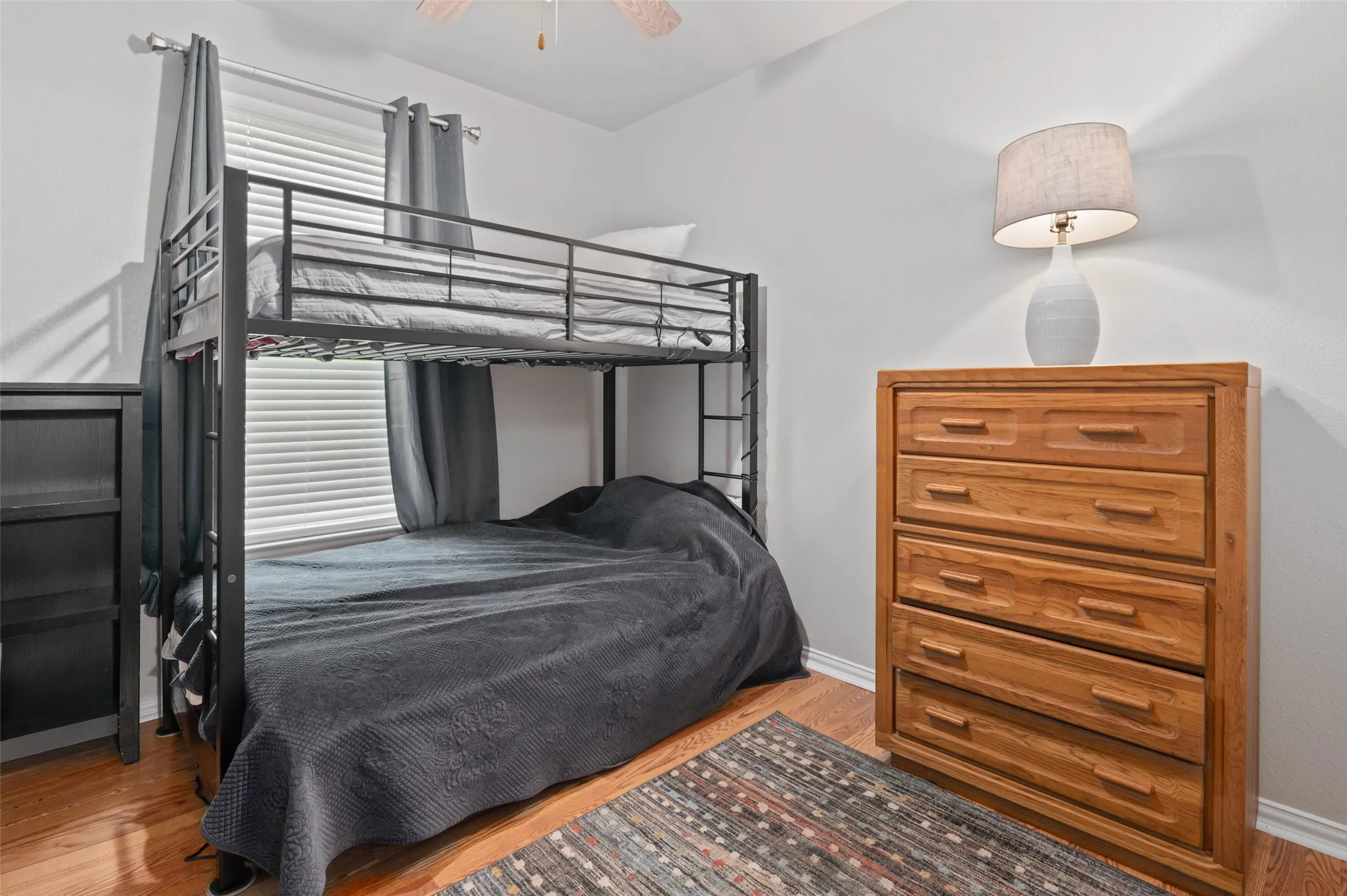 Bedroom featuring wood-finish flooring, white walls, and a window with blinds and gray drapes