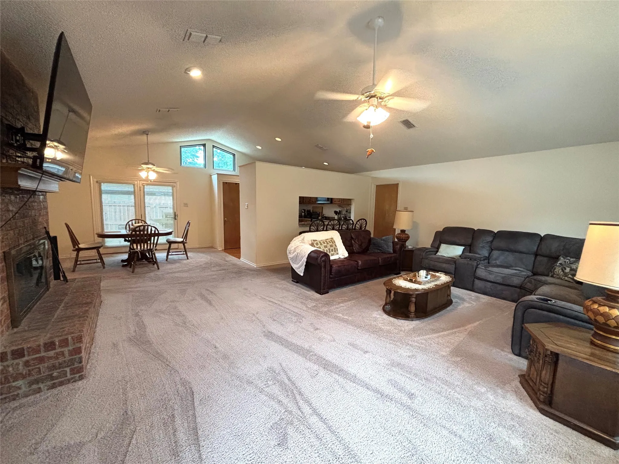 Spacious living area featuring light-toned carpeting, a brick fireplace with a hearth, and a vaulted ceiling