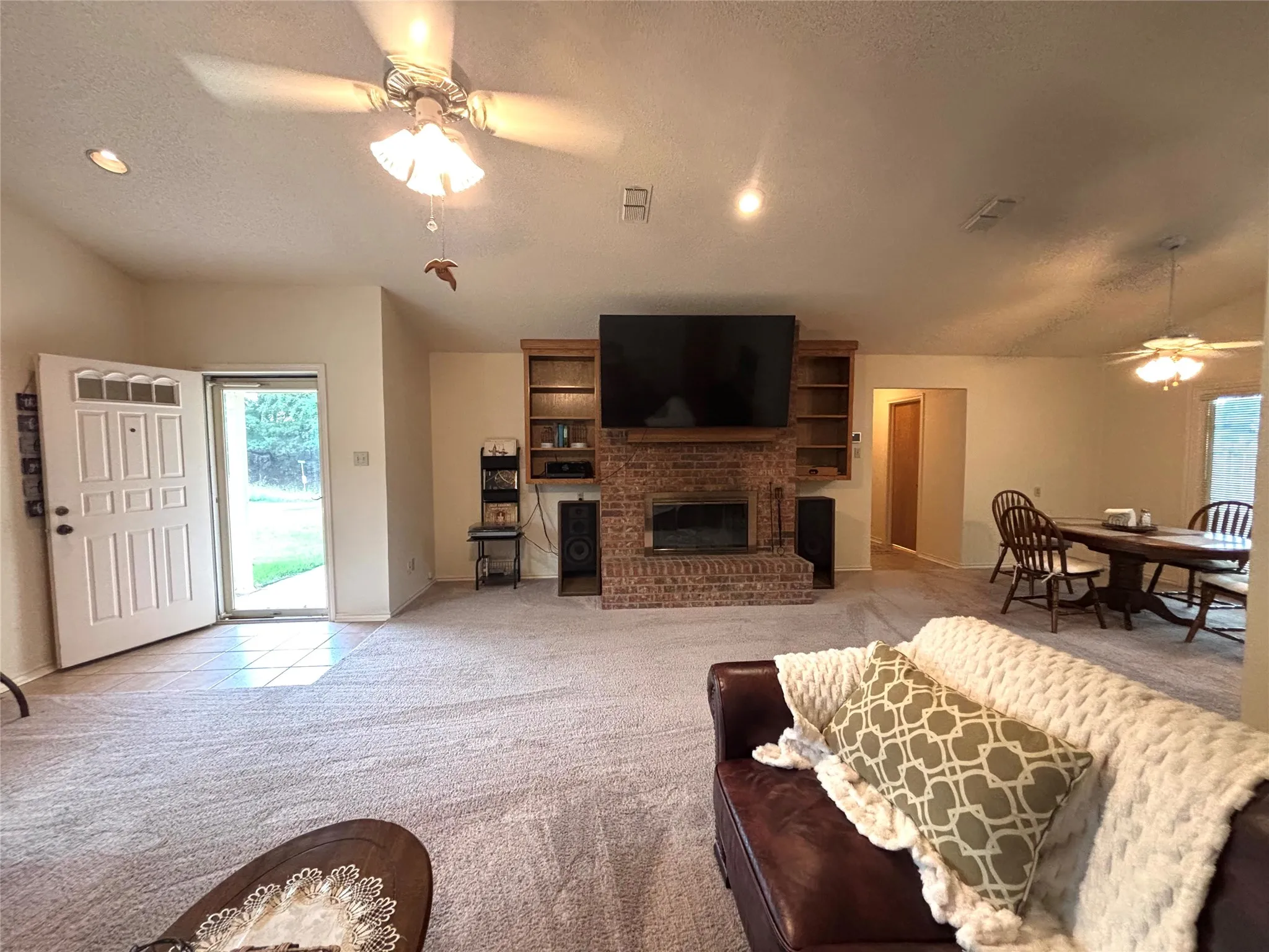 Spacious living area featuring a brick fireplace with wood-finish built-in shelving, vaulted ceilings, and light-toned carpeting
