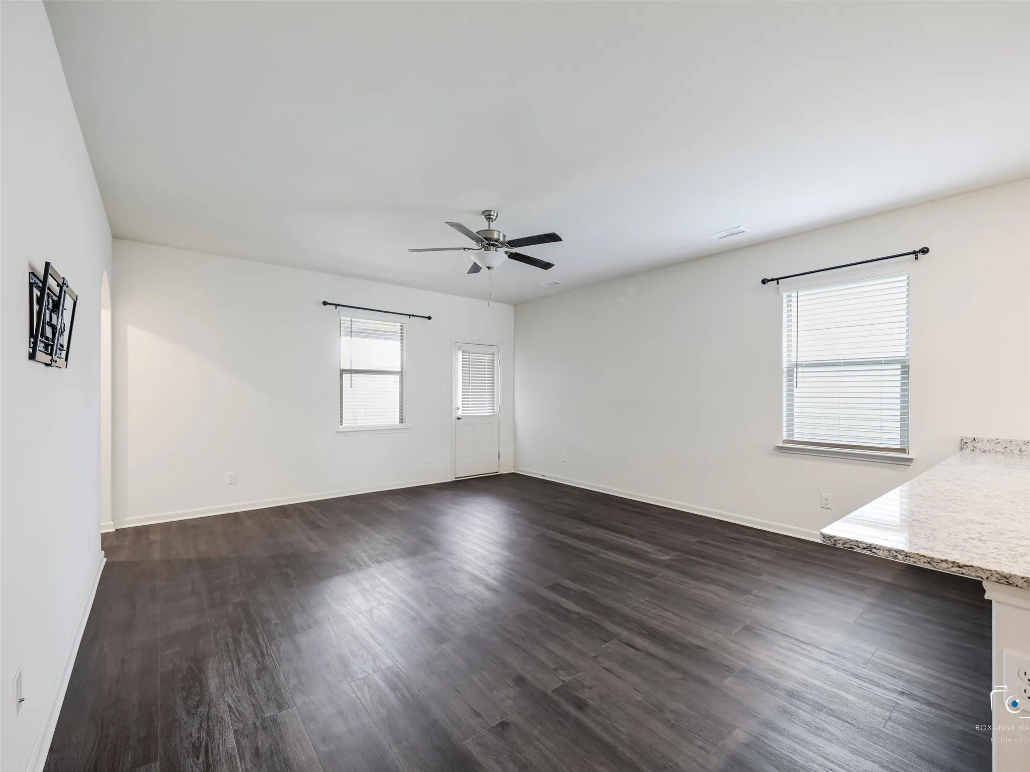 Spacious room featuring dark wood-finish flooring, white walls, two windows with blinds, a ceiling fan, and a granite countertop
