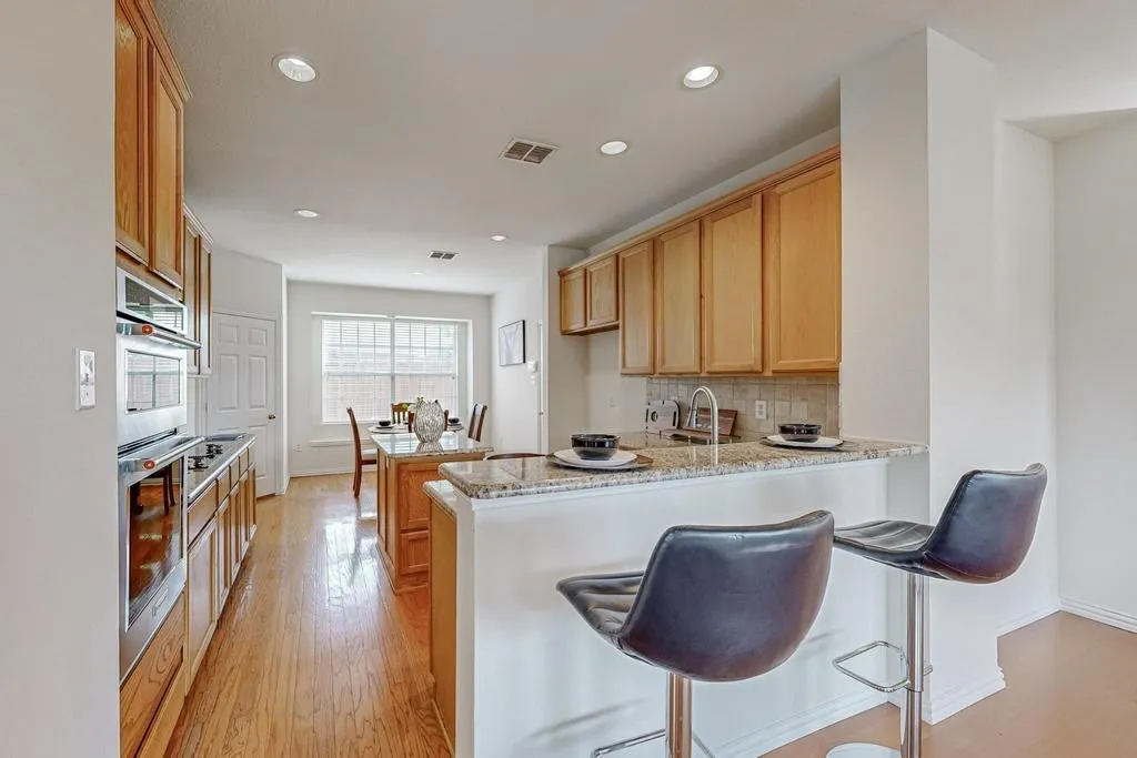 Kitchen featuring wood-finish flooring, light wood cabinetry, stainless steel appliances, a stone-finish countertop island with bar seating, and recessed lighting