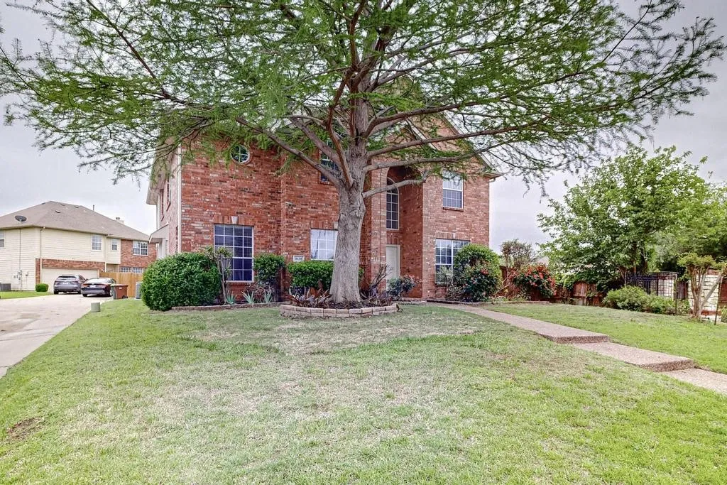 Two-story brick exterior with a prominent front-facing gable, mature front yard tree, established shrubbery, and a concrete walkway to the entrance
