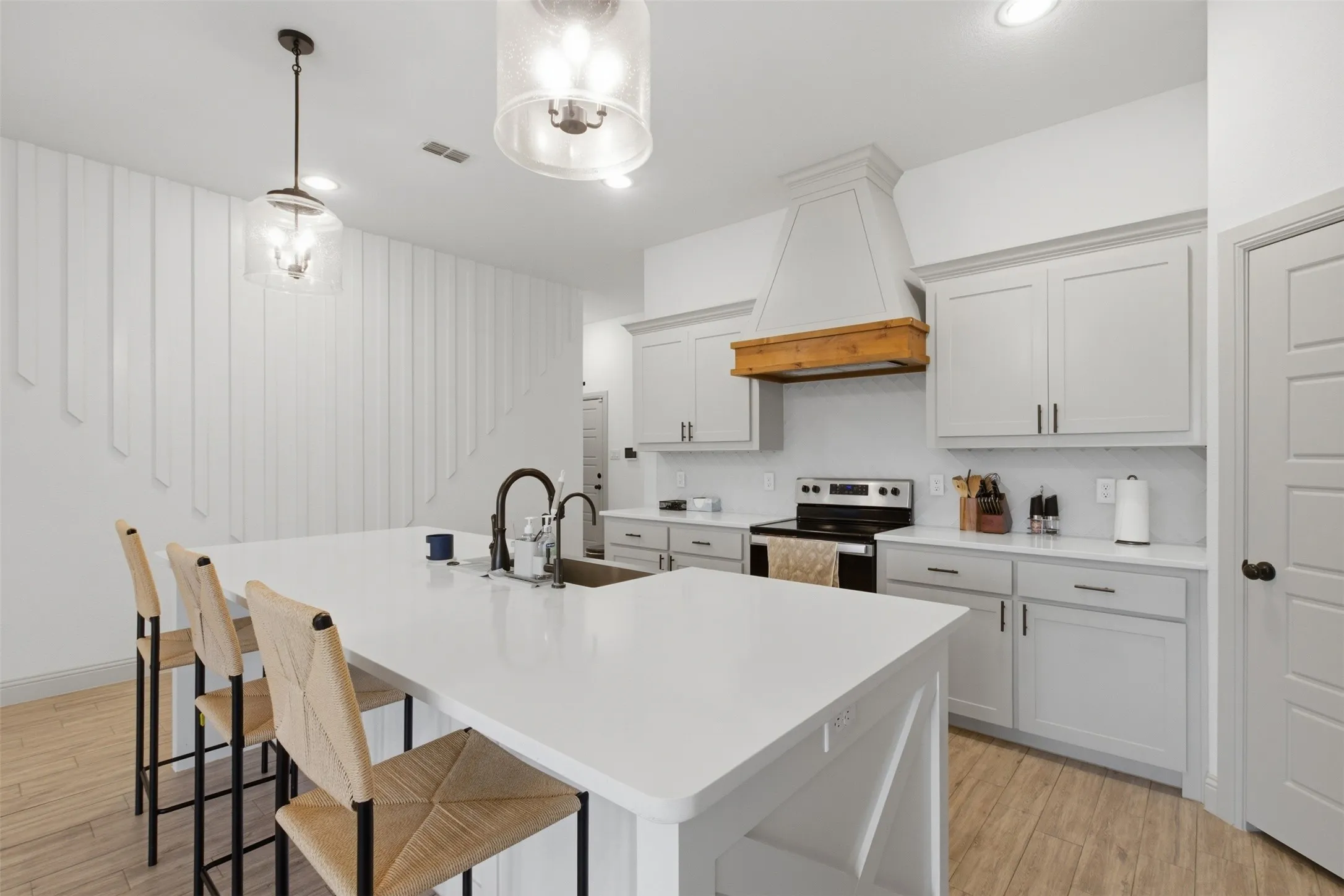 Spacious kitchen featuring a stainless farm style sink, island with integrated sink, white cabinetry with dark hardware, a built-in range hood with wood accent, and wood-finish flooring