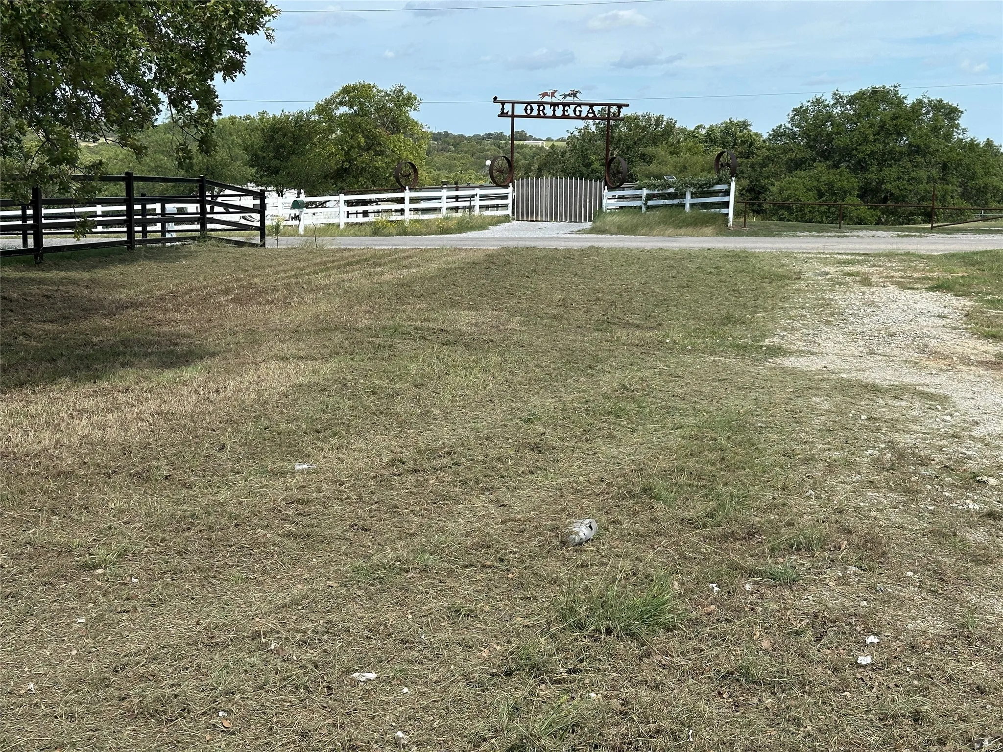 Gated entrance featuring a decorative metal archway, white perimeter fencing, and established trees