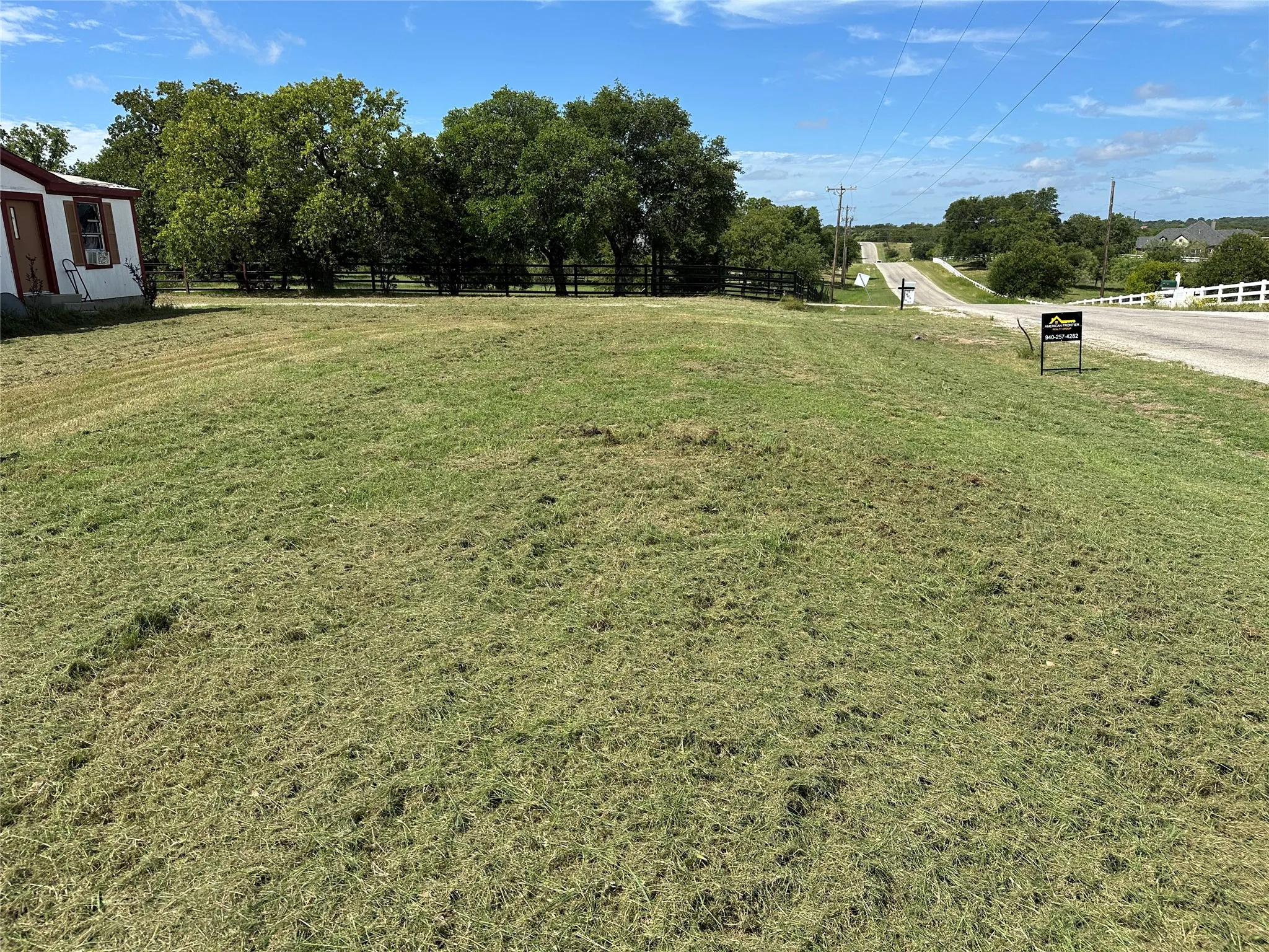 Expansive grassy lot featuring mature trees, a paved roadway, and a white split-rail fence