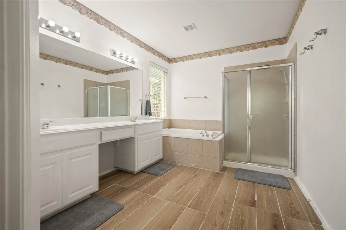 Spacious bathroom featuring wood-finish flooring, a dual-sink vanity with white cabinetry, a built-in bathtub with tiled surround, a separate glass-enclosed shower, and a large mirror with overhead lighting