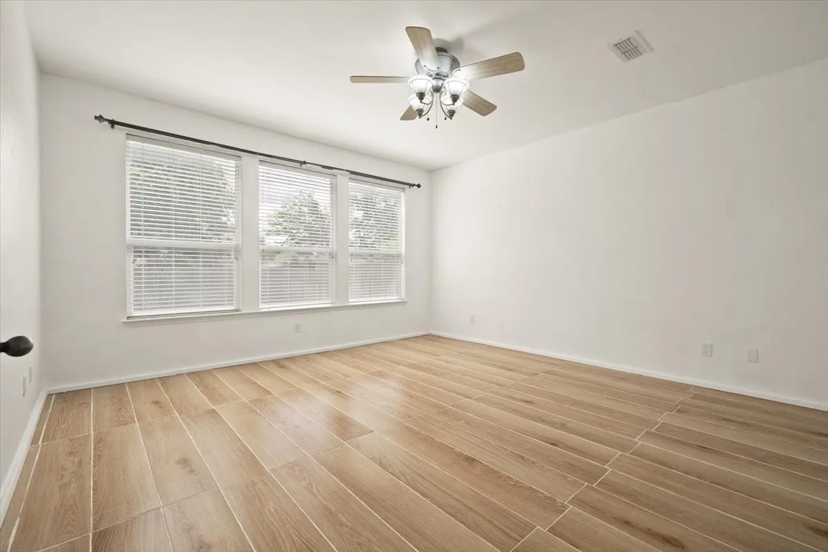 Spacious room featuring wood-finish flooring, three large windows with blinds, a ceiling fan with integrated lighting, and white baseboards