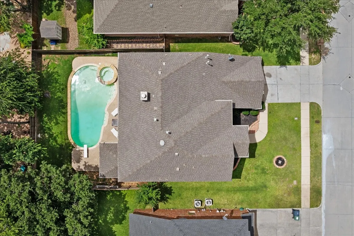 Aerial property overview featuring a private swimming pool with integrated spa, a concrete pool deck, green lawn areas, mature trees, and a shingle roof