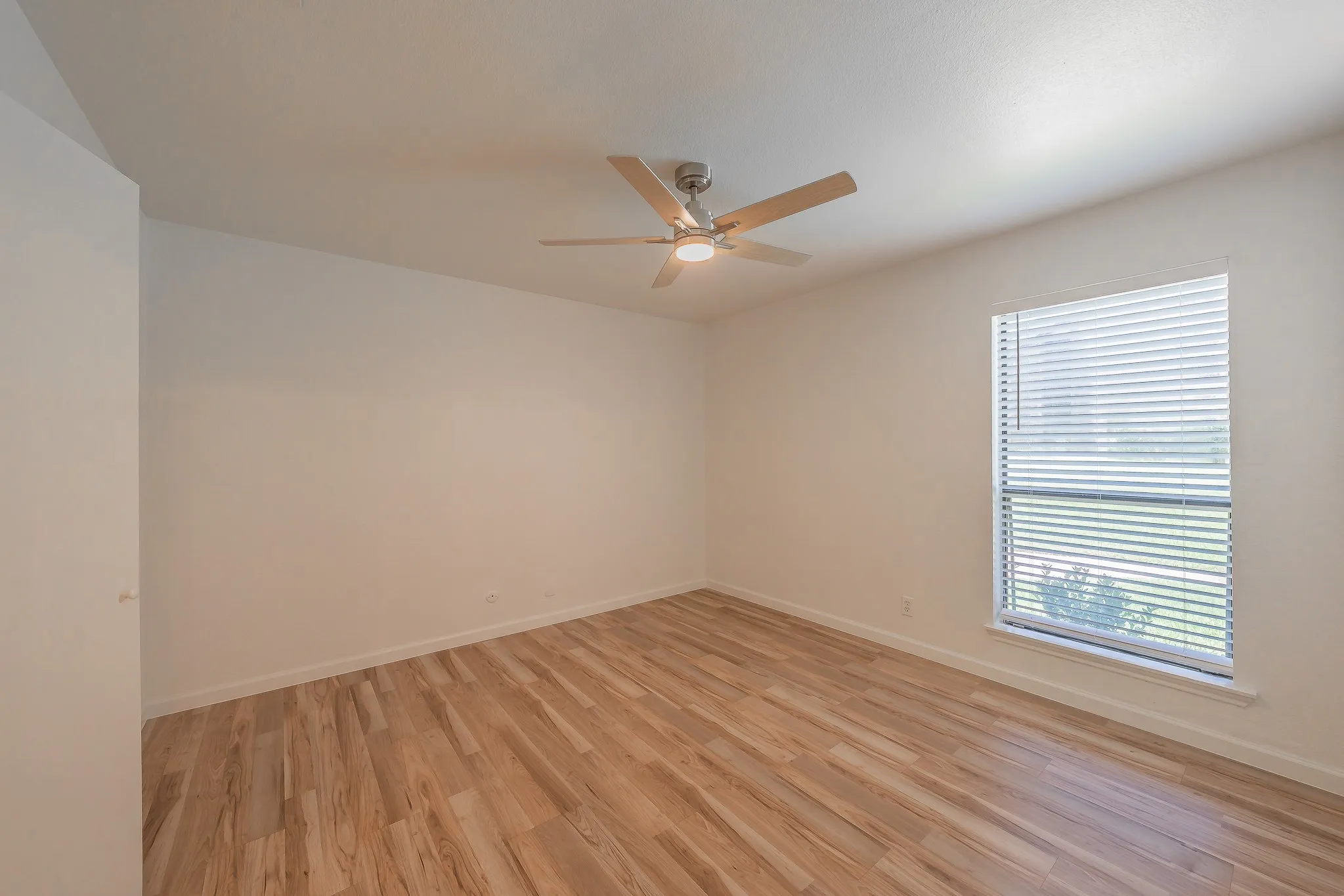 Spacious primary bedroom featuring light wood-finish flooring, a new contemporary ceiling fan with integrated lighting, white walls, and a window equipped with blinds