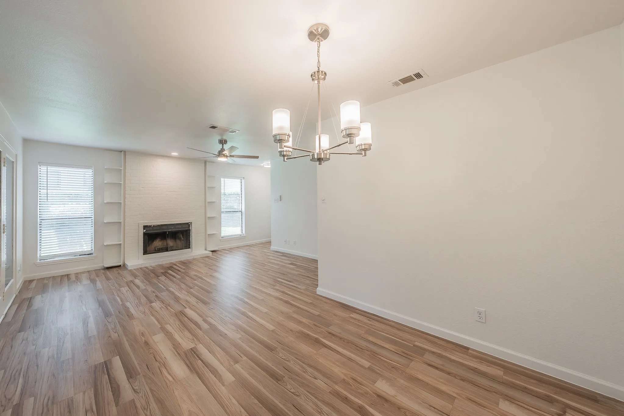Open-concept living space featuring wood-finish flooring, a white brick fireplace with built-in shelving, and a contemporary chandelier