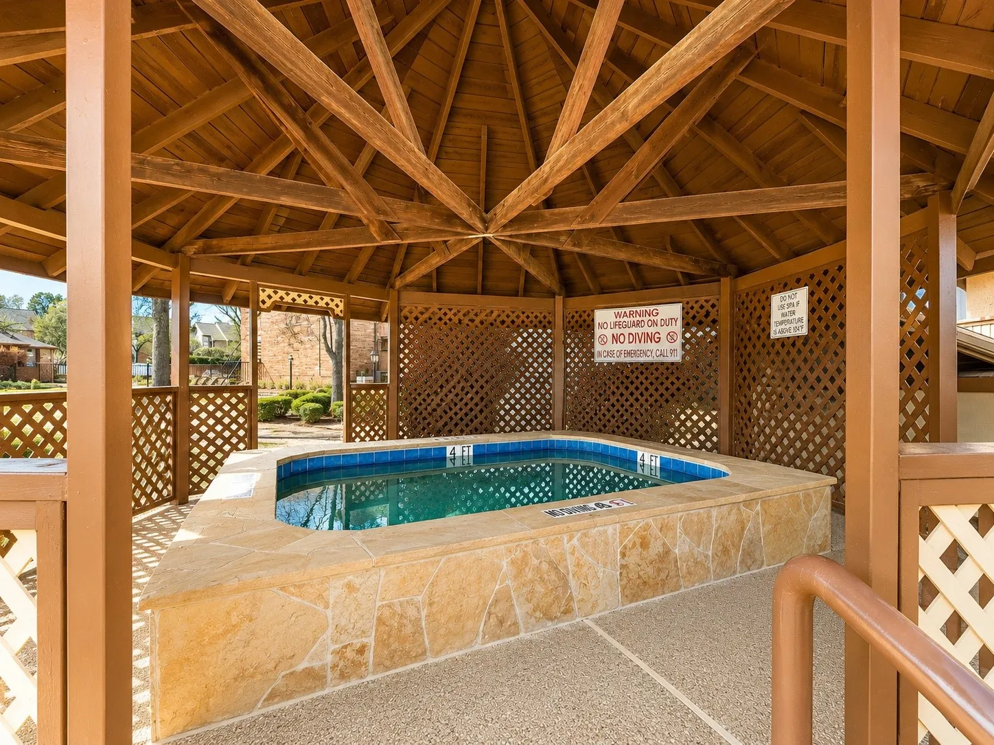 Gazebo-covered spa with stone surround and blue tile accents, featuring a wood lattice enclosure and exposed aggregate flooring