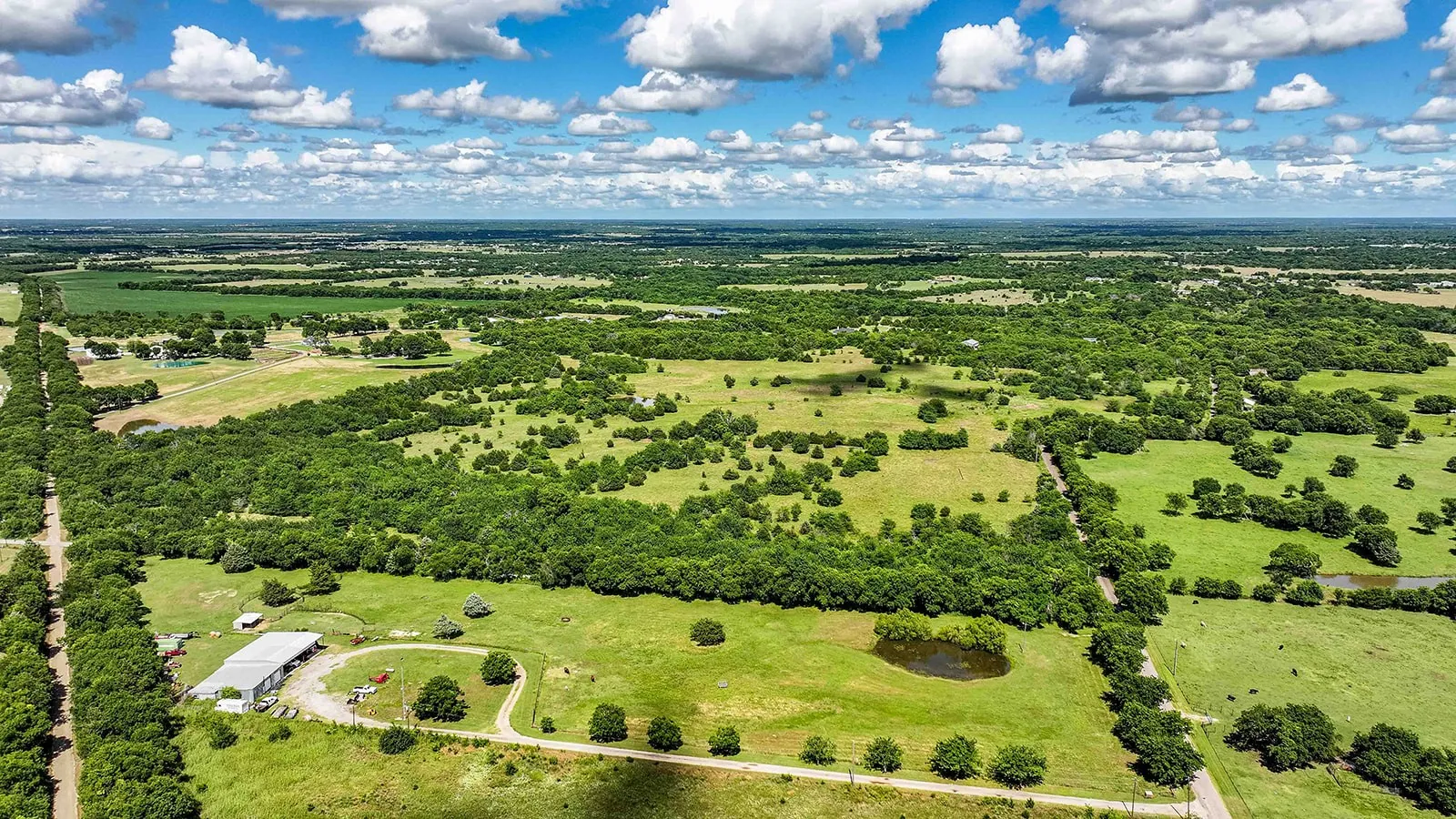 Expansive rural property featuring a large metal-roofed barn, an unpaved circular drive, a small pond, and abundant mature trees