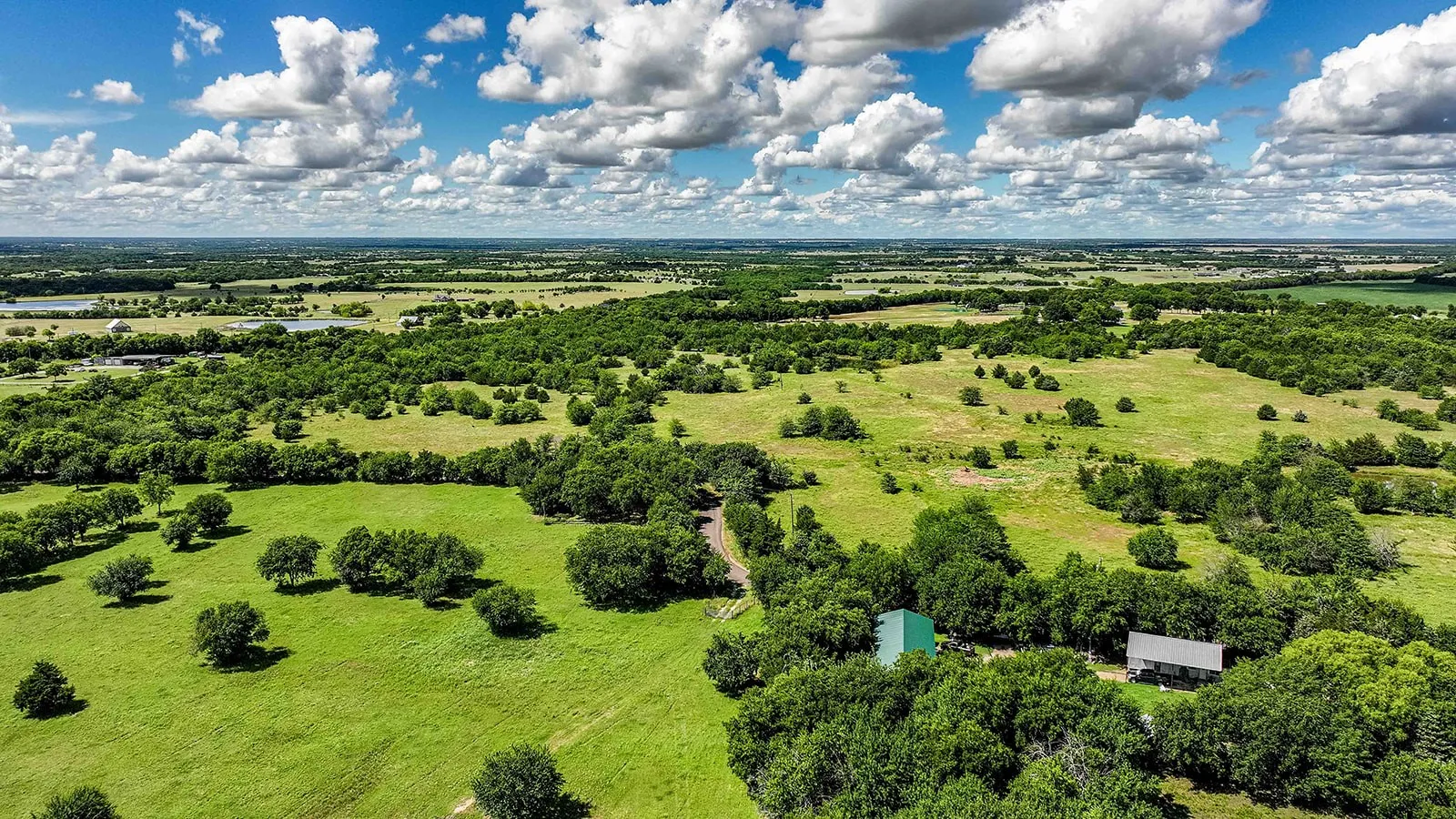 Expansive green landscape featuring scattered trees, a winding dirt path, and two structures with pitched roofs