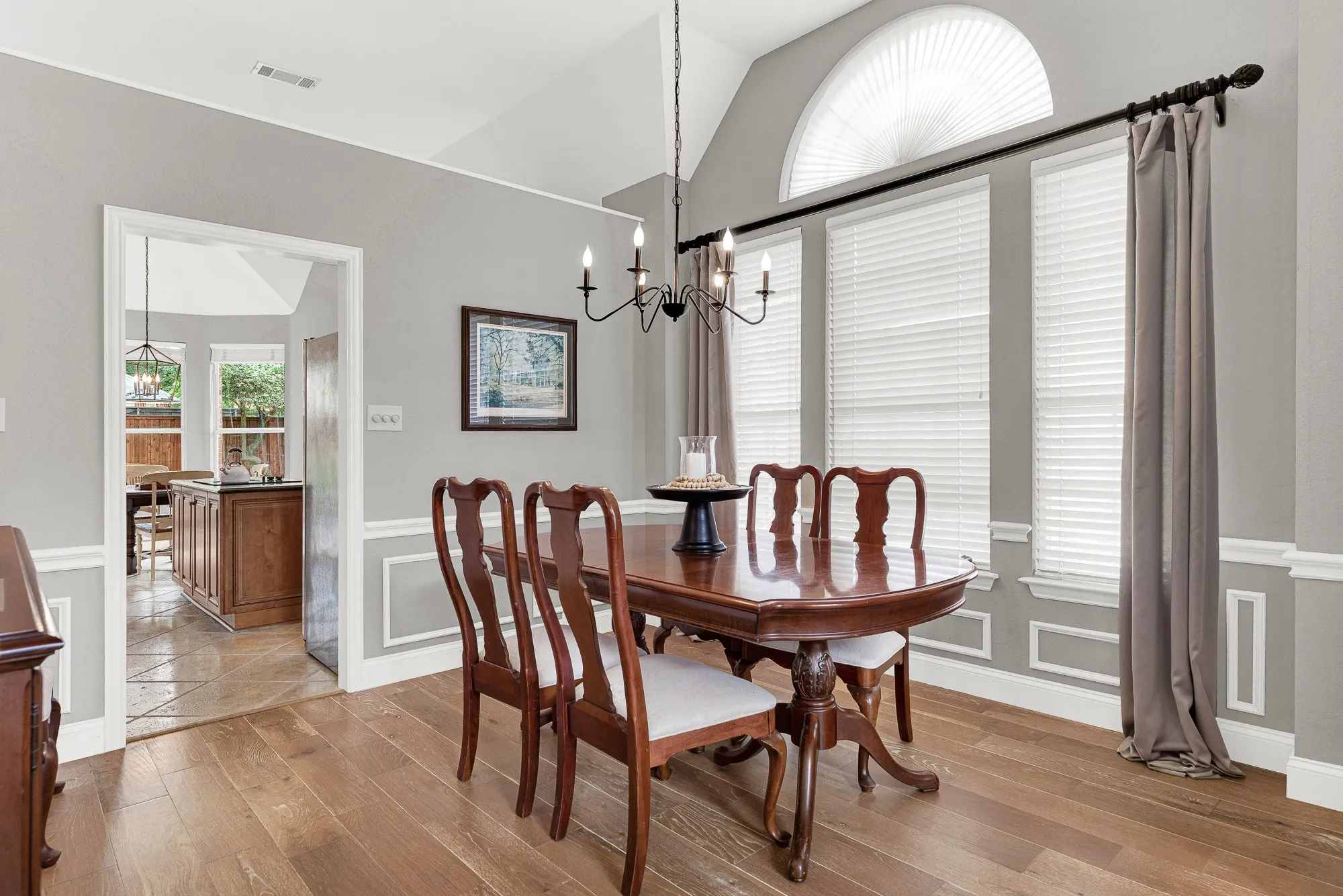 Vaulted ceilings rise dramatically above picture-frame wainscoting, while a tall arched transom window draws natural light deep into the space from two directions. This room was made for a proper dinner party — the kind where nobody checks their phone