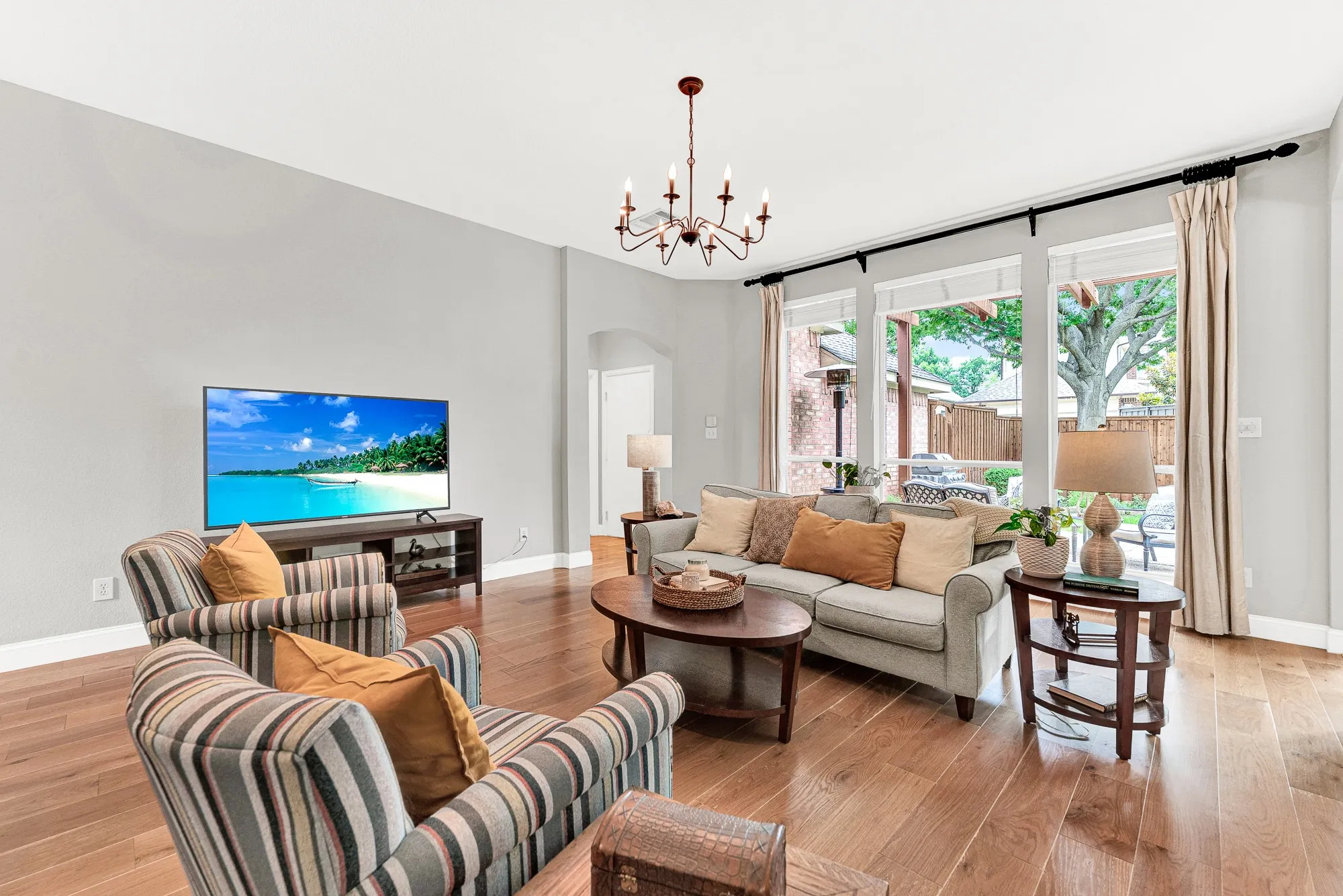 Two oversized windows with floor-length panels frame a leafy backyard view, flooding the wide-plank hardwood floors with warm natural light from the pergola and mature oak tree beyond. On a bright afternoon, the inside and outside feel like one continuous living space.
