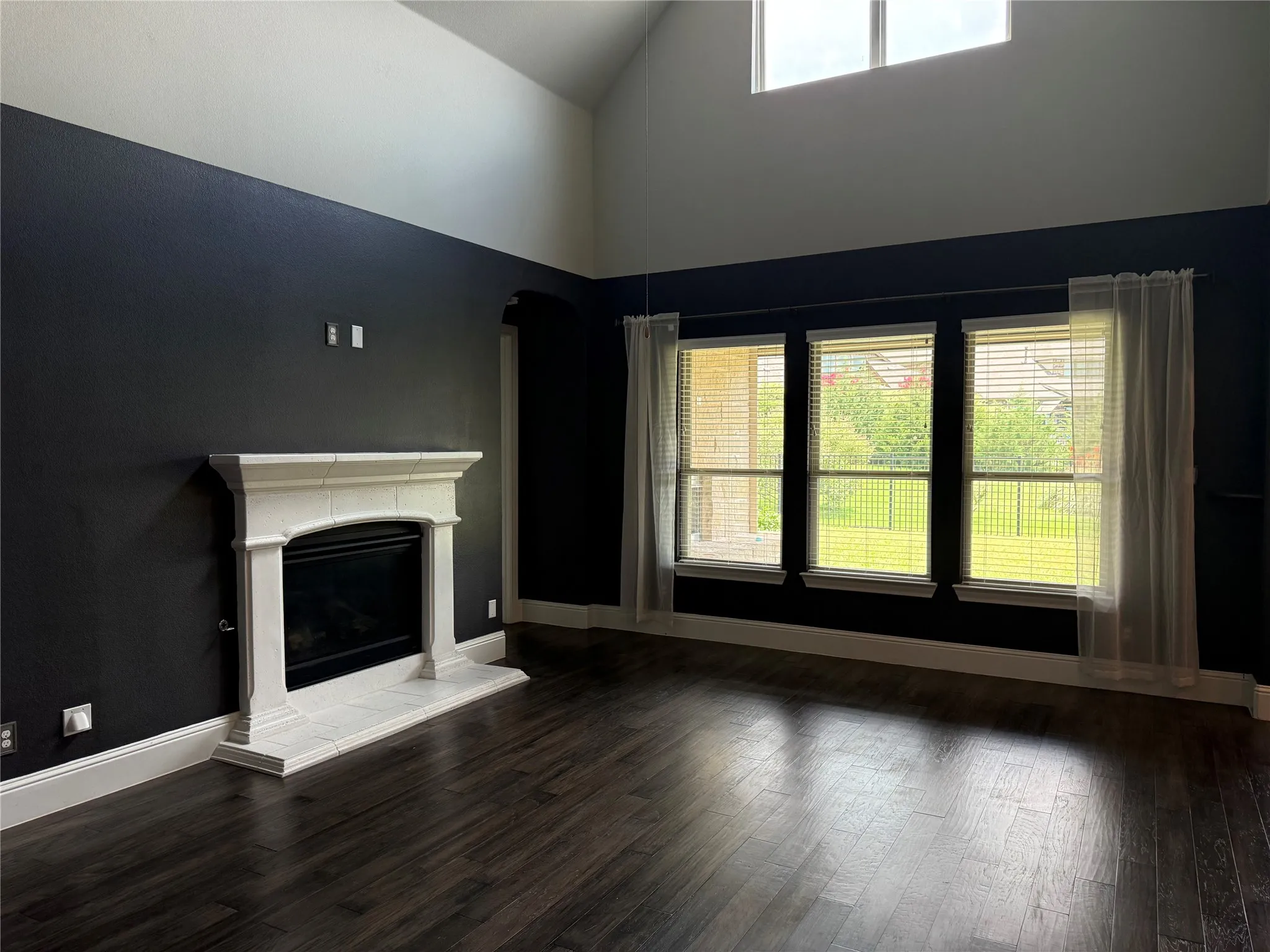 Vaulted ceiling living space featuring a white mantel fireplace, dark wood-finish flooring, and multiple windows with blinds