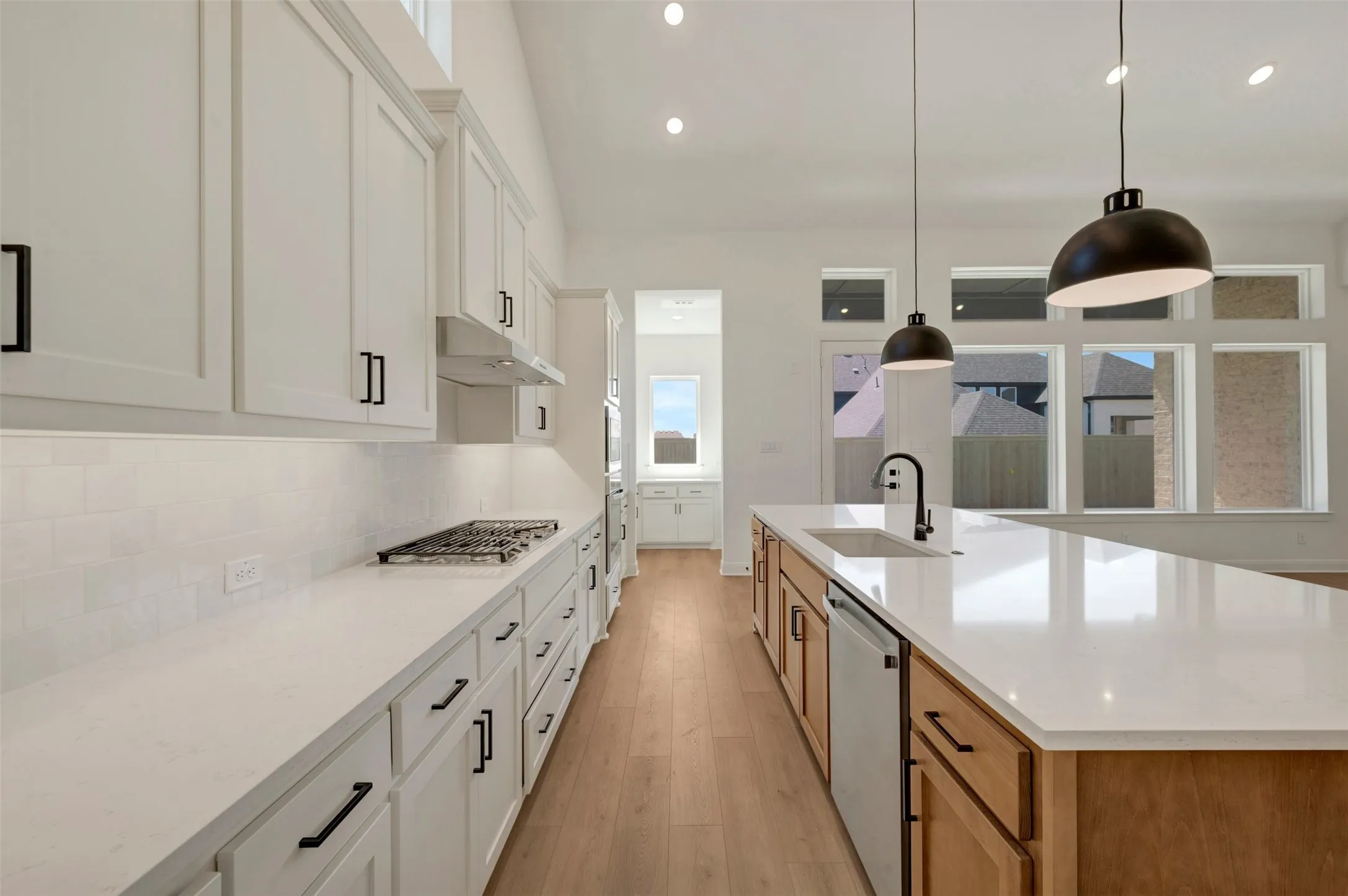 Vaulted ceiling with recessed lighting, wood-finish flooring, white cabinetry with matte black hardware, white subway tile backsplash, and an expansive kitchen island with a built-in sink and pendant lighting