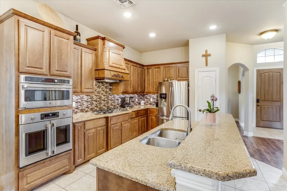 Kitchen featuring extensive wood cabinetry, stainless steel appliances, a built-in oven and microwave, a multi-toned tile backsplash, and a large island with an undermount dual-basin sink and a gooseneck faucet