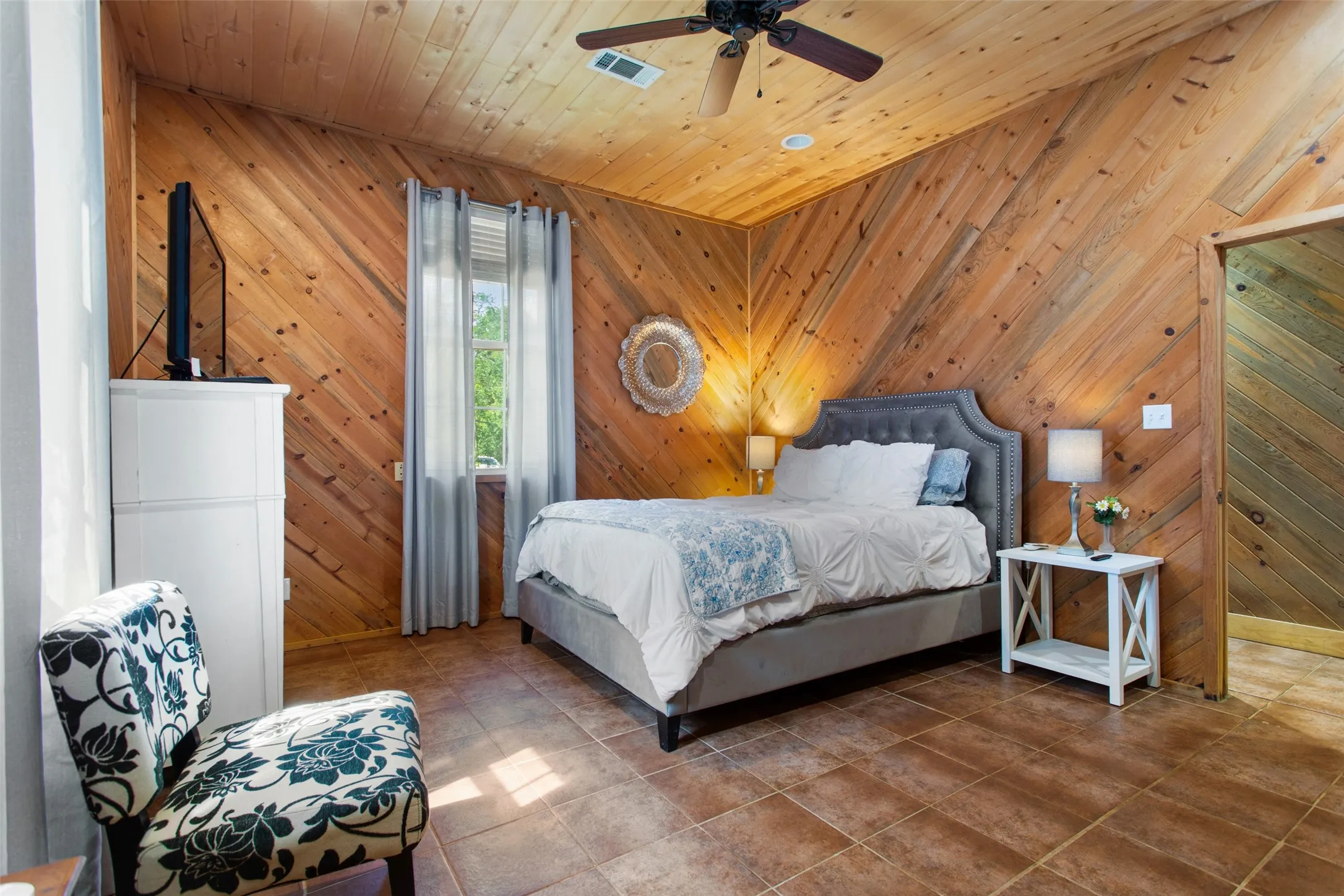 Room featuring angled wood paneling on walls and ceiling, brown tile flooring, and a window with sheer and blackout curtains