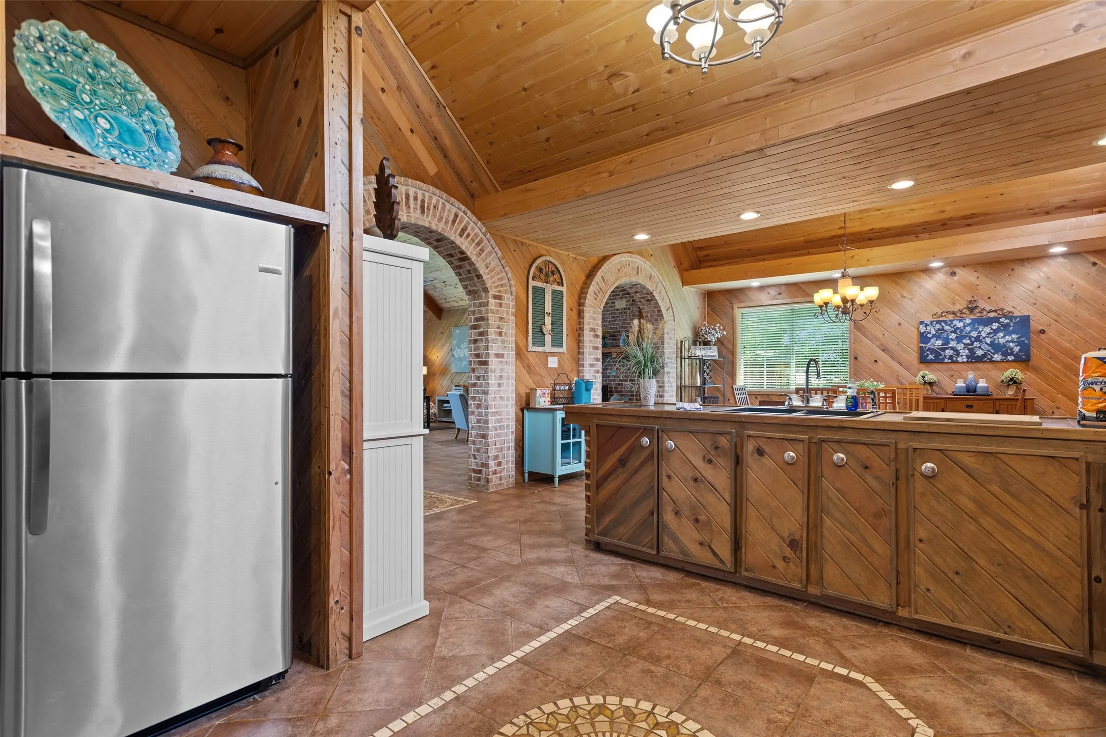 Kitchen featuring wood-paneled walls and ceiling, custom wood cabinetry, stainless steel refrigerator, and tiled flooring with decorative inlay