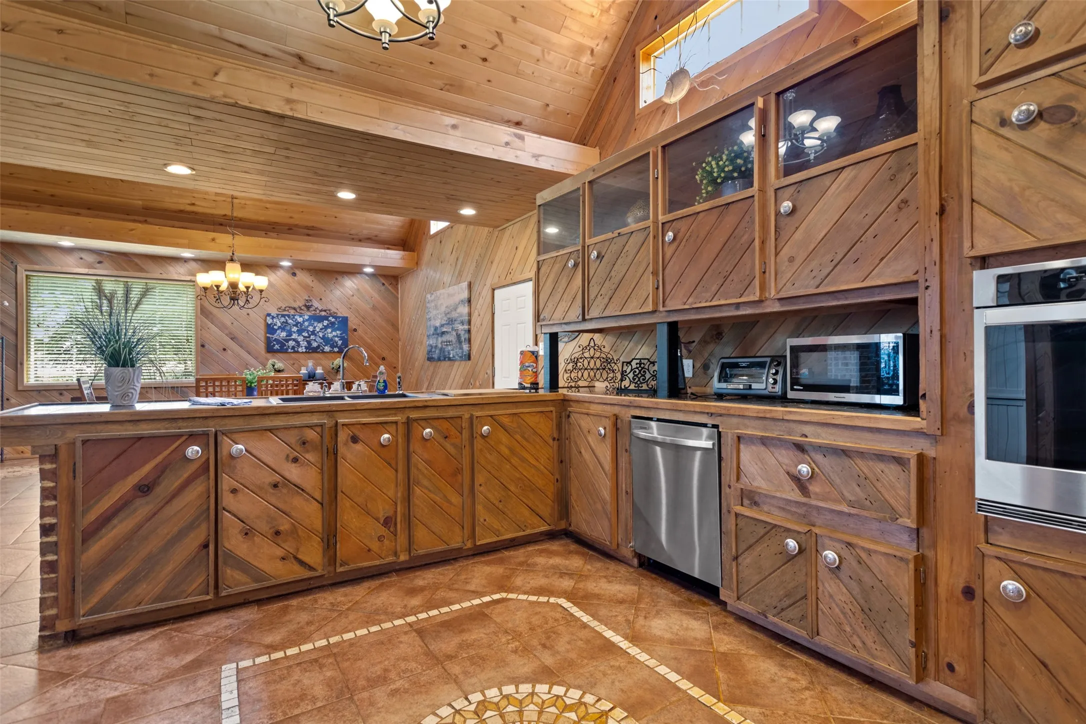 Kitchen featuring extensive wood cabinetry, stainless steel appliances, tile flooring with mosaic details, and a vaulted wood-paneled ceiling