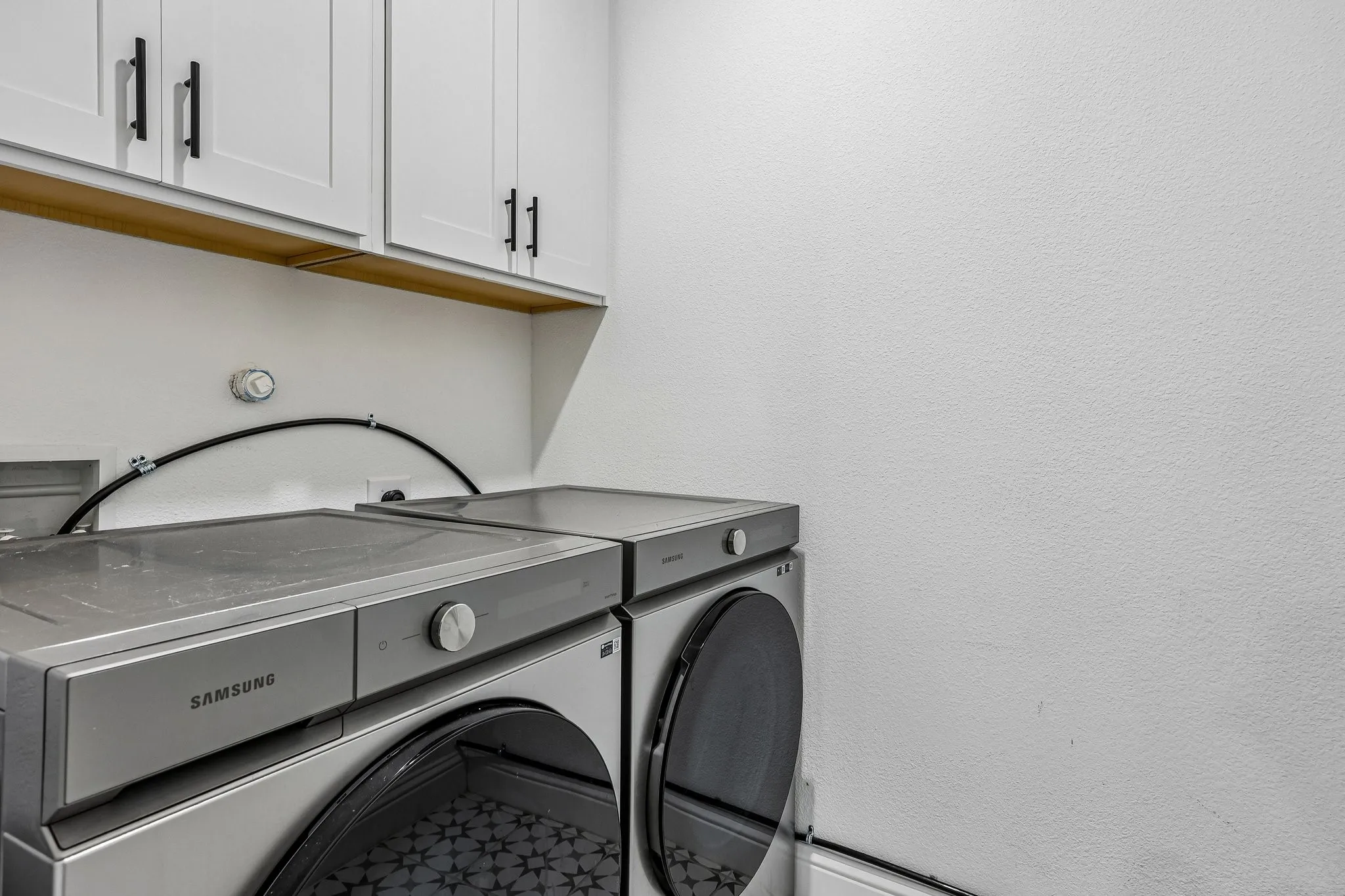 Dedicated laundry area featuring white upper cabinetry with black hardware, a light wood-finish shelf, and patterned floor tile