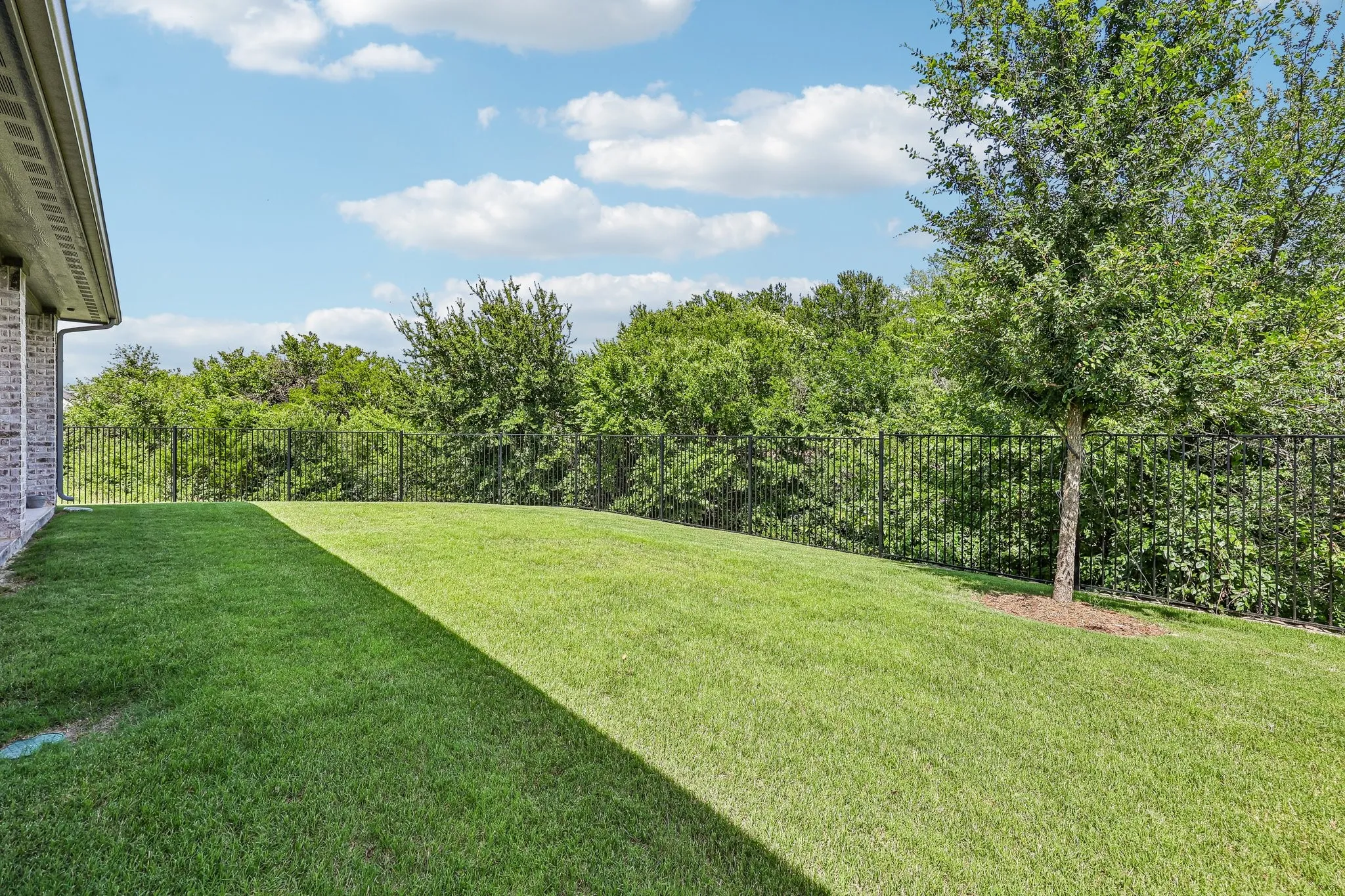 Expansive lawn area featuring a black metal fence, mature trees, and a brick exterior wall
