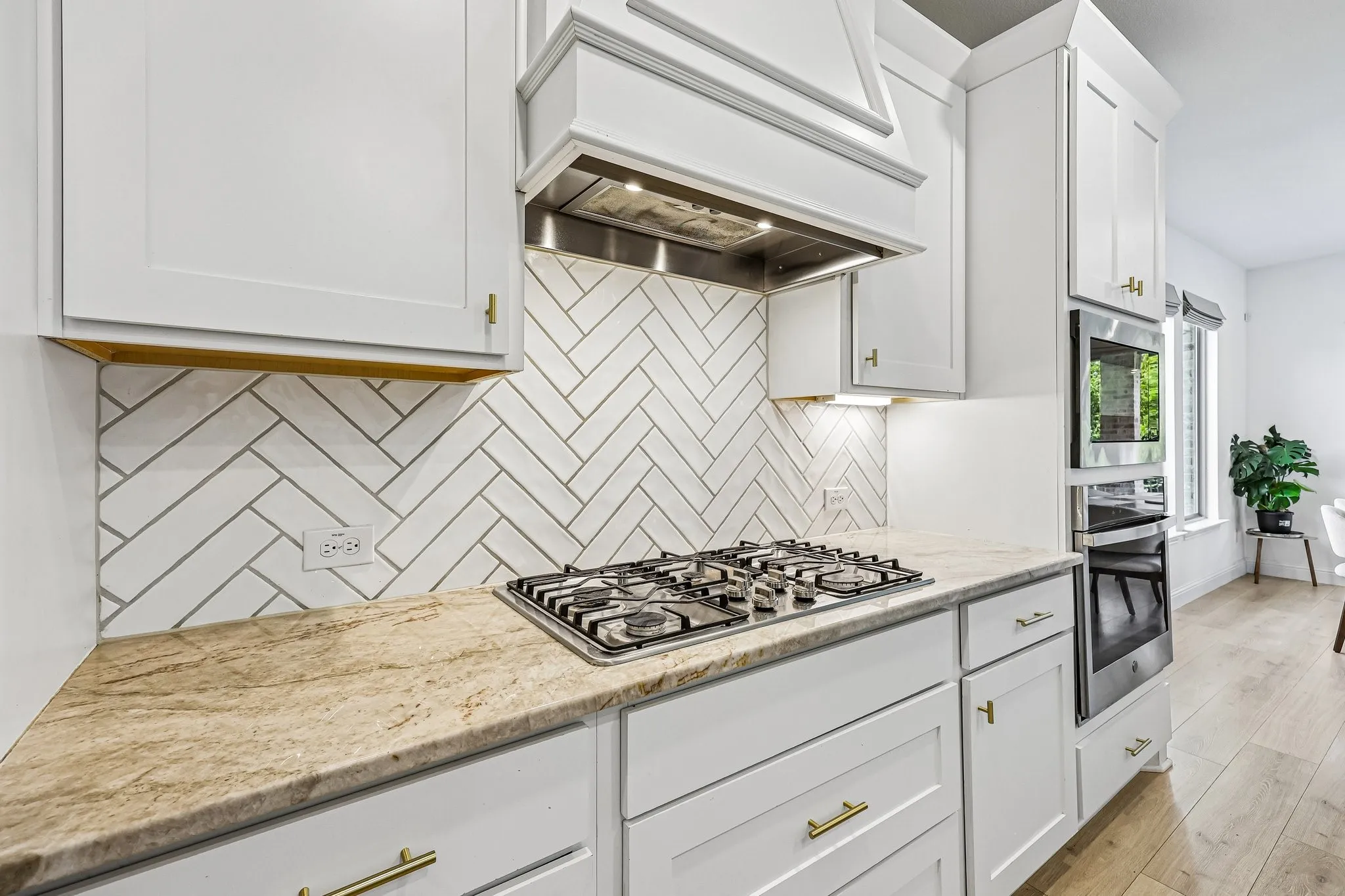 Modern kitchen featuring a gas range with stainless steel hood, built-in double ovens, light-toned countertops, white cabinetry with gold-toned hardware, and a herringbone pattern tile backsplash