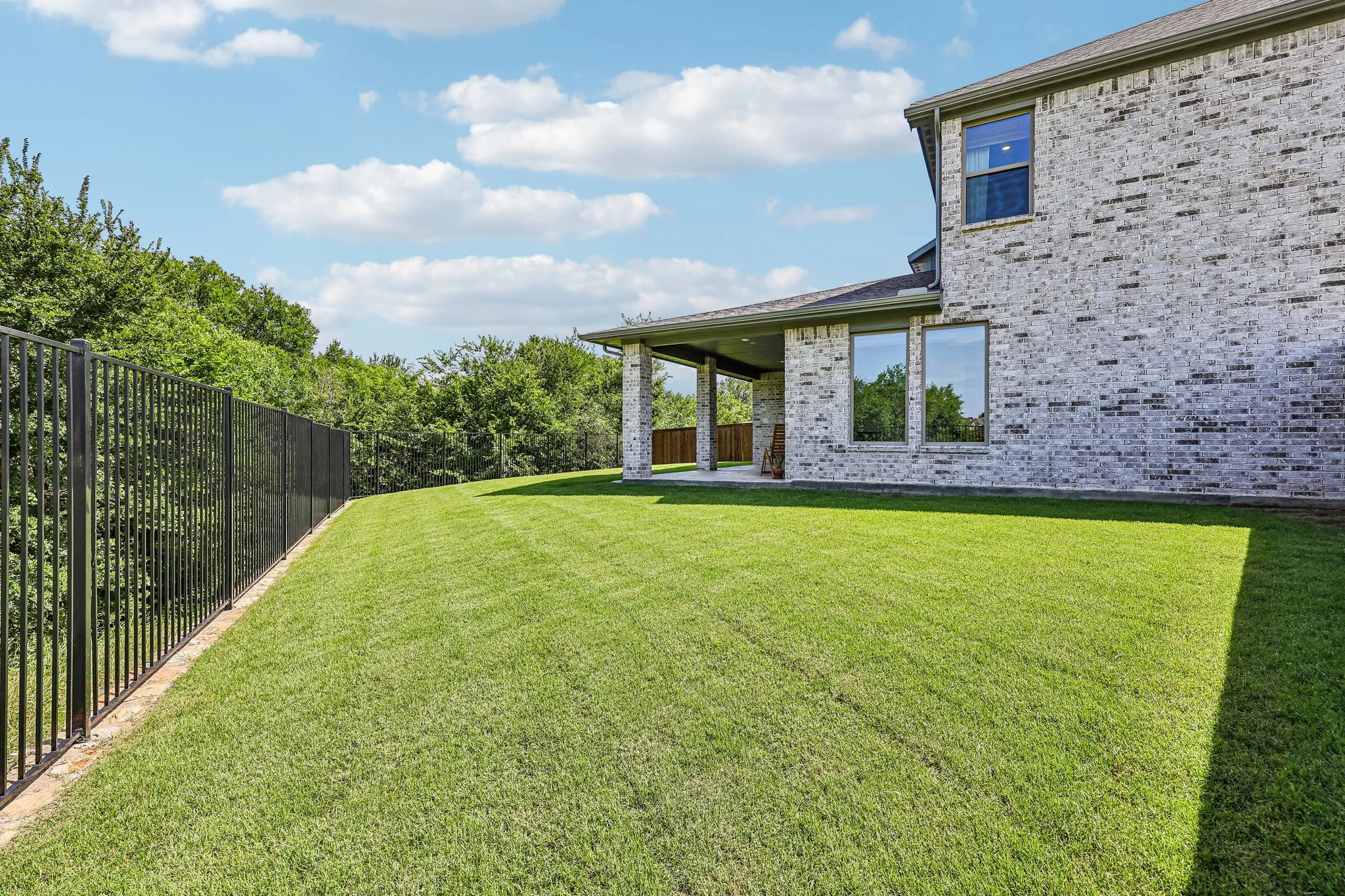 Expansive green space featuring a multi-story brick exterior, covered patio with column accents, large windows, and a black metal fence bordering mature trees