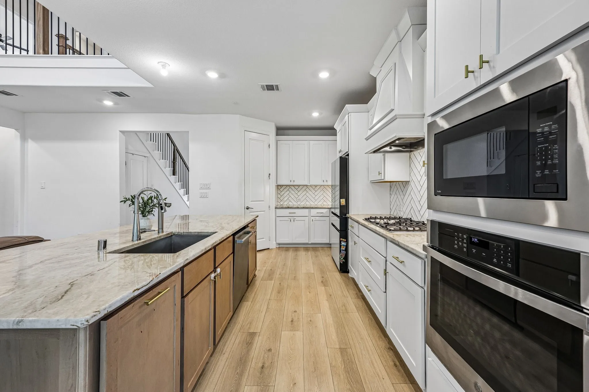 Kitchen featuring light wood-finish flooring, white cabinetry with gold-tone hardware, a large island with an undermount sink, and stainless steel appliances including a built-in microwave and oven