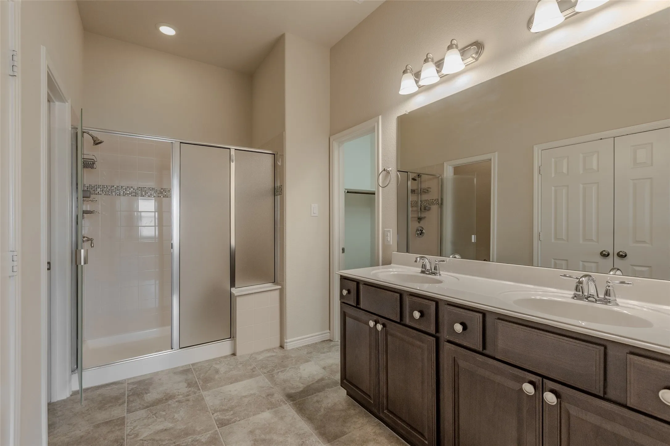 Bathroom featuring a dual vanity with white countertops, a large frameless mirror, and a shower enclosure with a tiled accent band