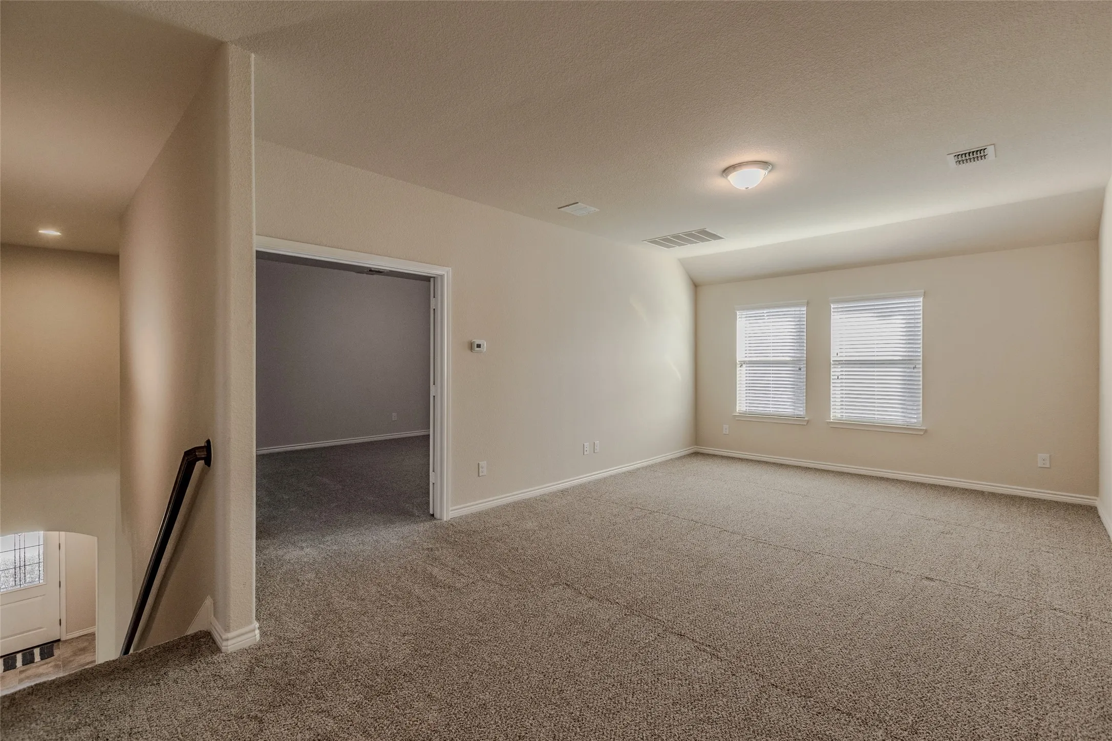Spacious carpeted upstairs room featuring two windows with blinds, a ceiling-mounted light fixture, and a doorway opening to another room
