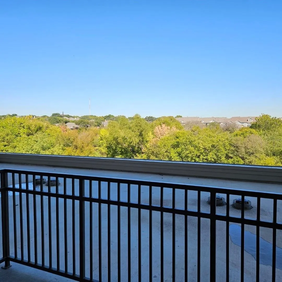Balcony with metal railing and concrete flooring, featuring a distant treeline and residential structures under a clear sky