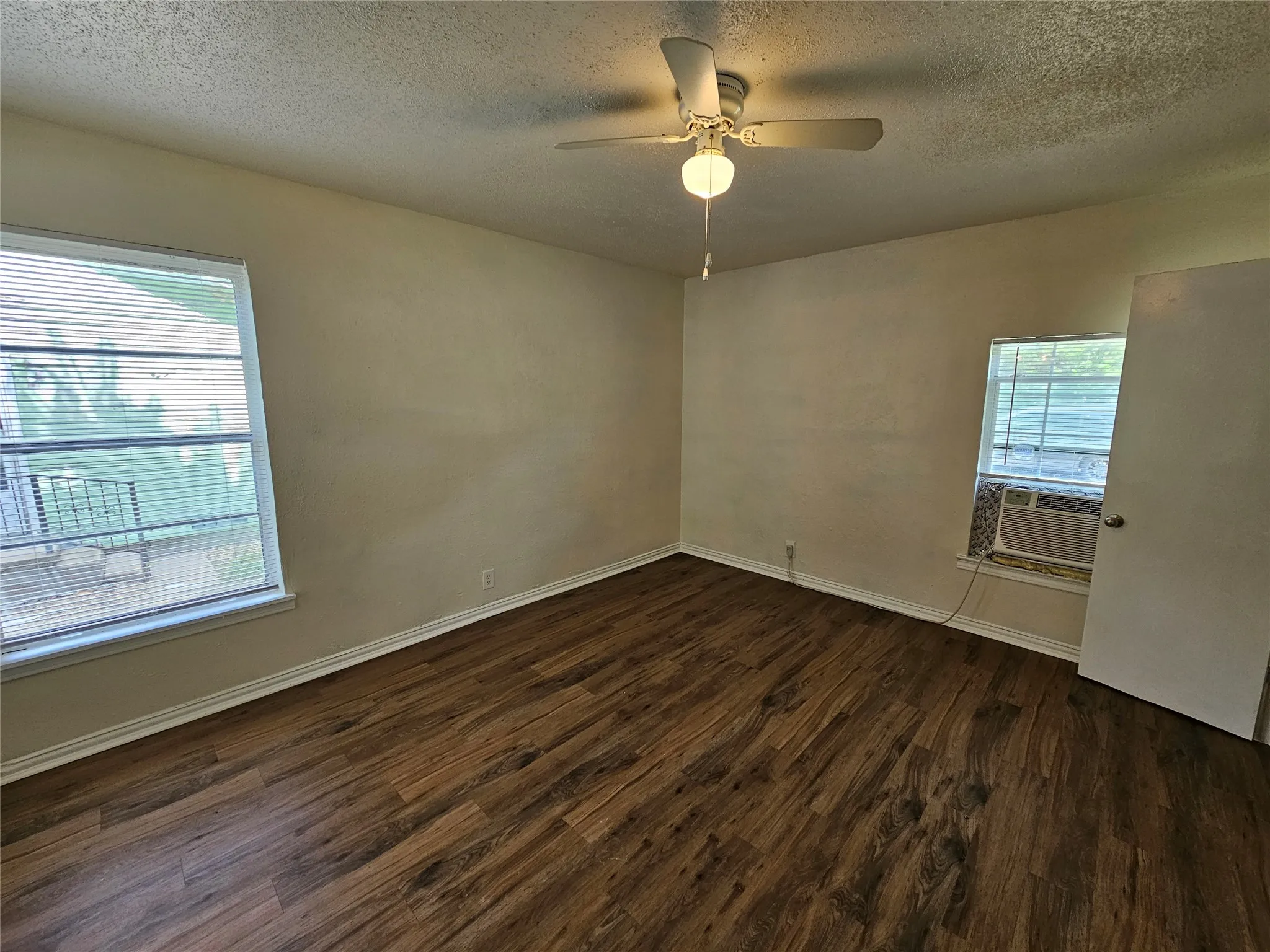 Spacious room with wood-finish flooring, two large windows with blinds, a ceiling fan with integrated lighting, and a wall-mounted air conditioning unit