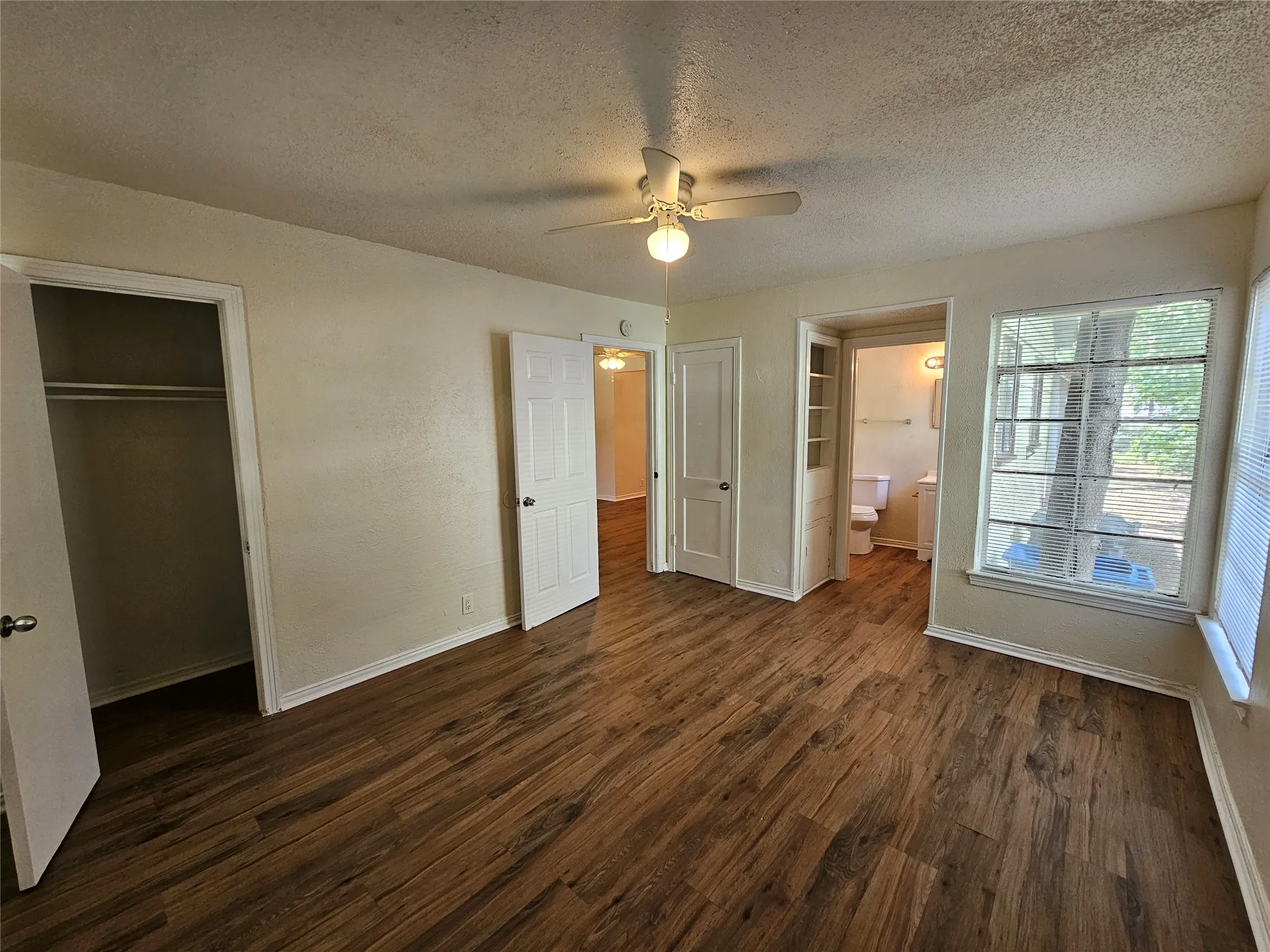 Spacious interior room featuring wood-finish flooring, a ceiling fan with integrated lighting, and multiple windows with blinds