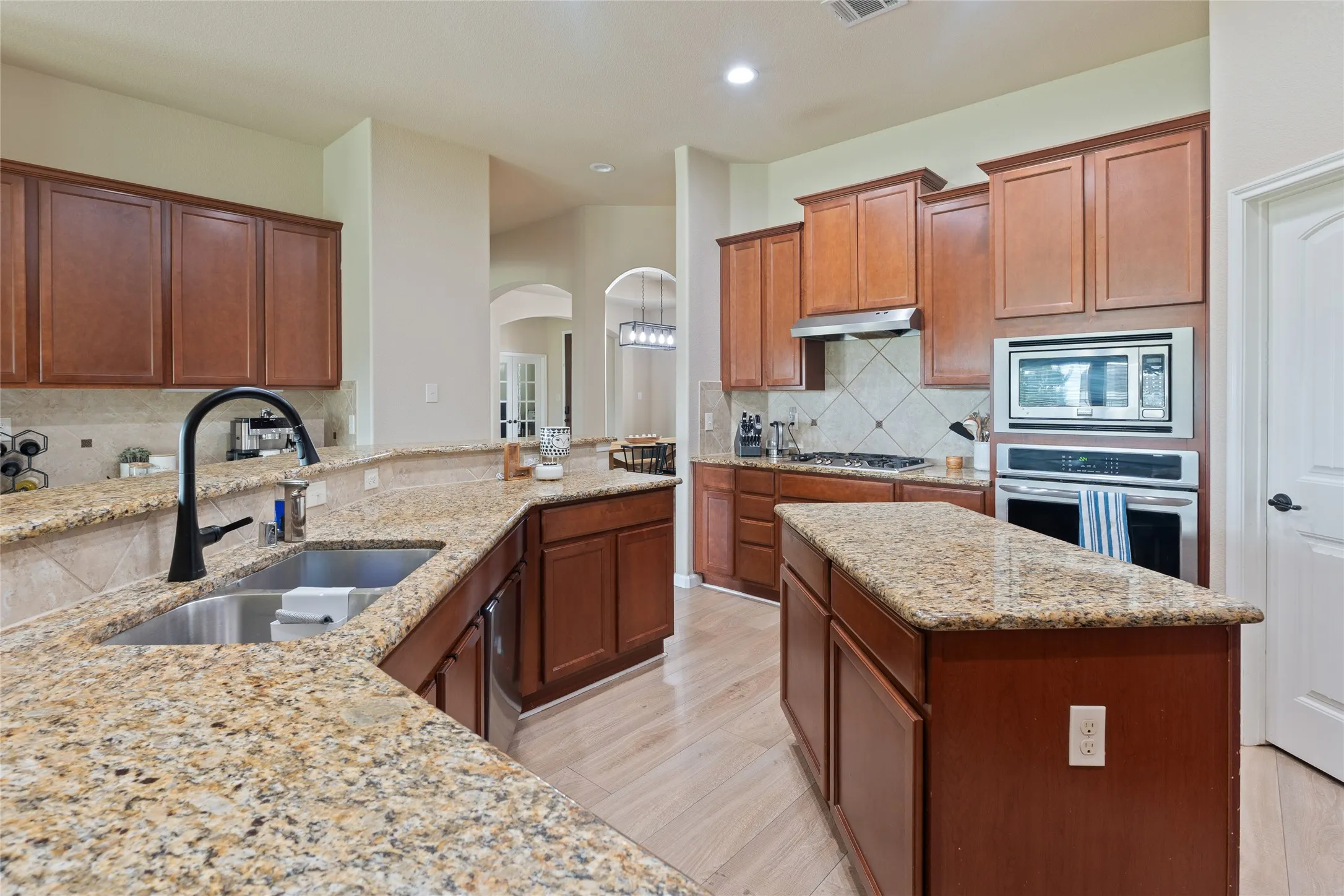 Kitchen featuring granite countertops, wood cabinetry, a double basin stainless steel sink with a matte black faucet, built-in stainless steel oven and microwave, and wood-finish flooring