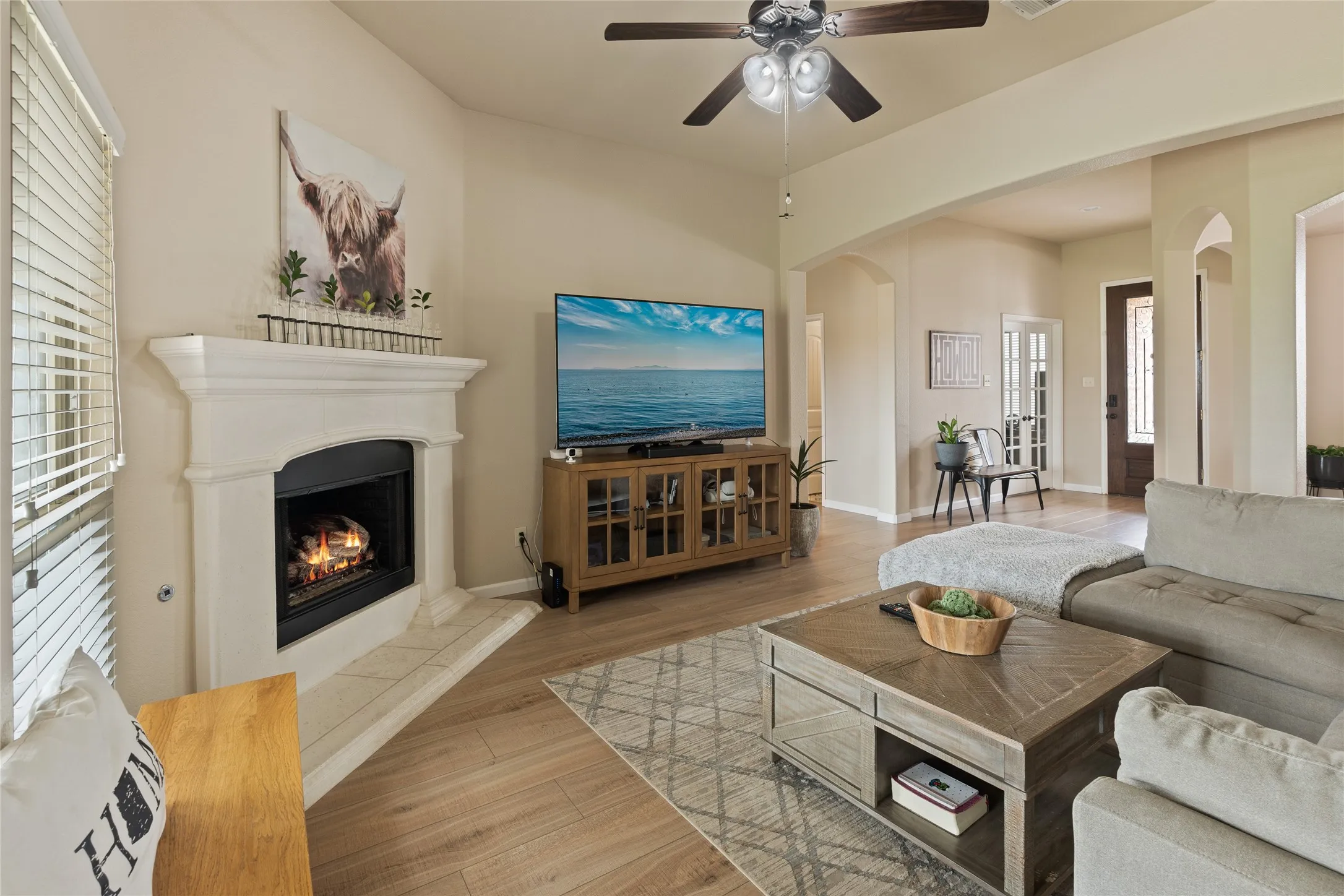 Living space featuring wood-finish flooring, a corner fireplace with a white mantel and hearth, a ceiling fan, and arched doorways