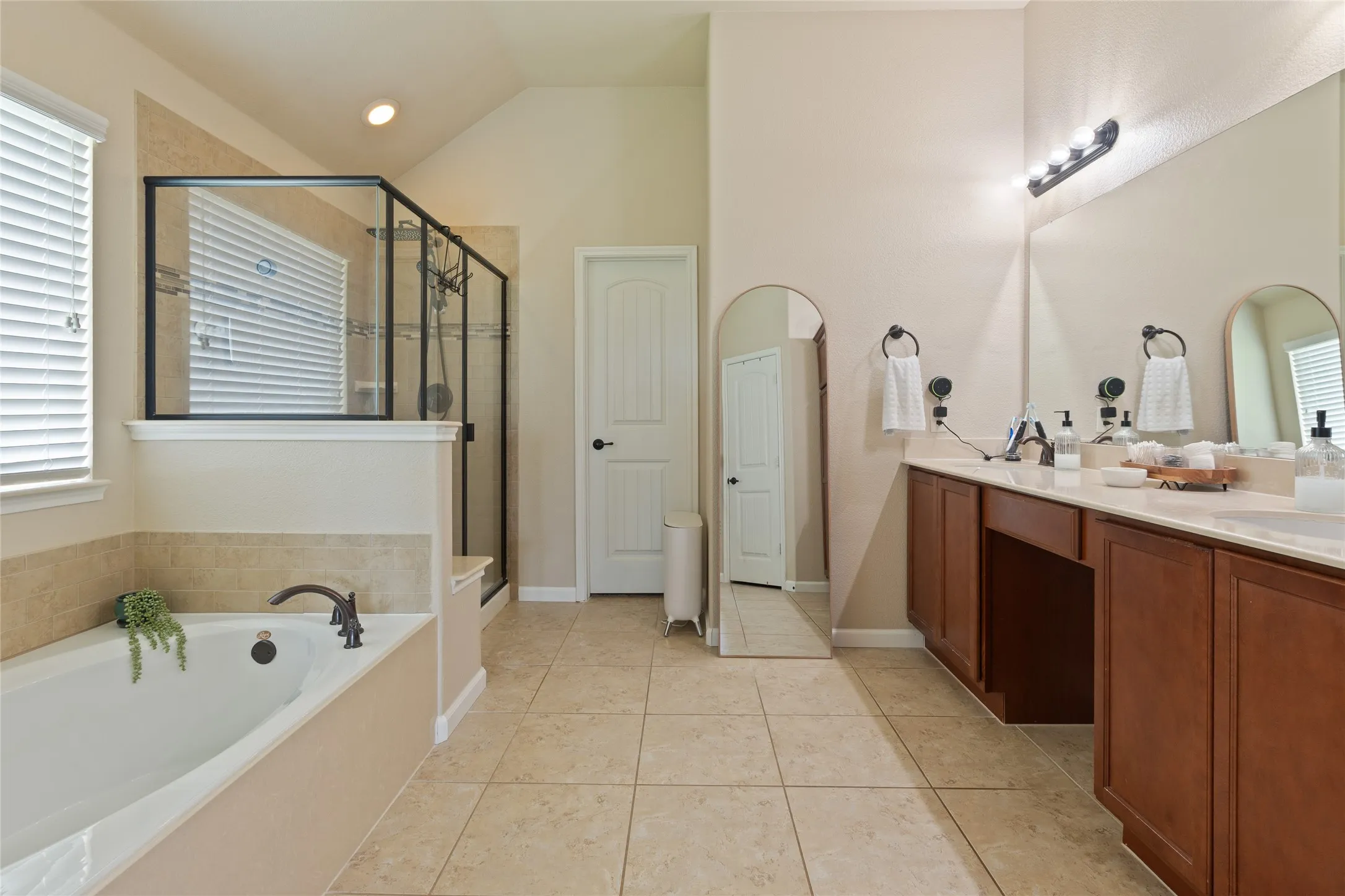 Spacious bathroom featuring a built-in soaking tub with a bronze-finish faucet, a glass-enclosed shower, a dual vanity with light-colored countertops, and tile flooring
