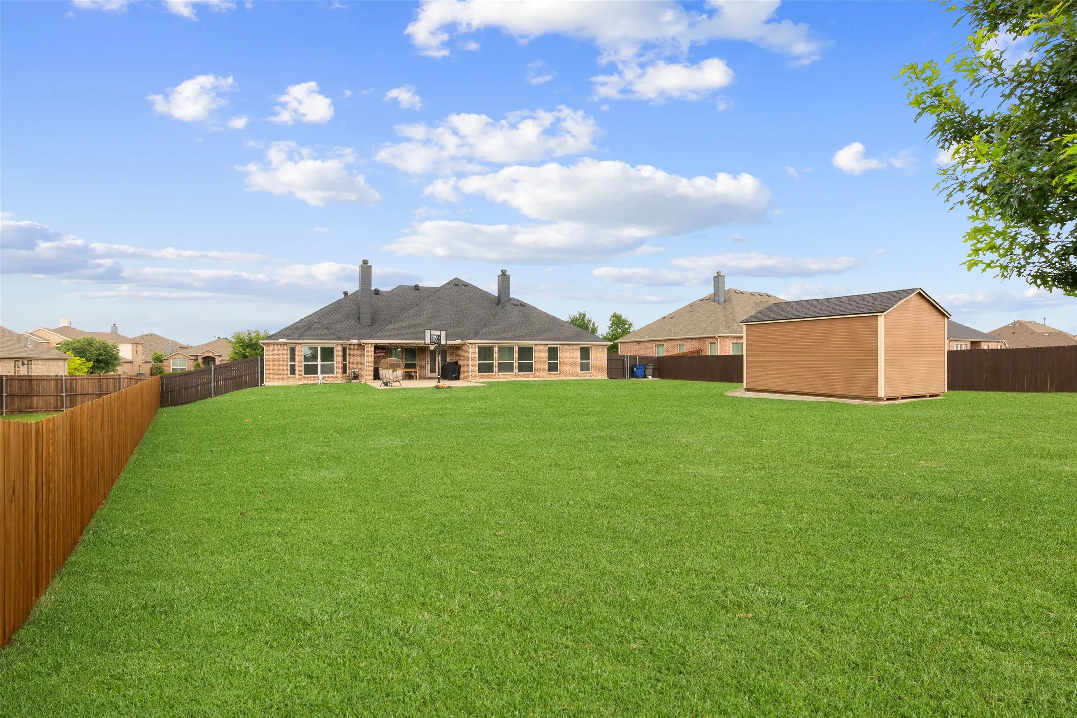 Expansive rear lawn enclosed by wood fencing, featuring a detached storage shed with a shingled roof, a paved patio, and a brick-clad exterior with multiple windows