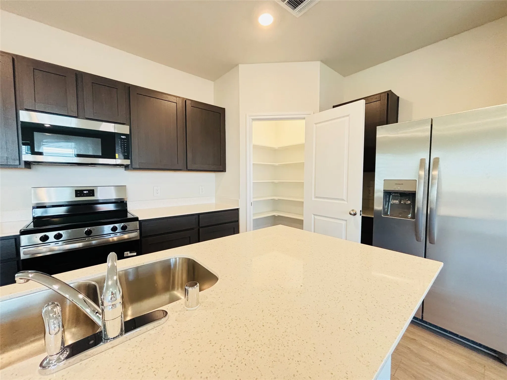 Modern kitchen featuring dark wood-finish cabinetry, stainless steel appliances, light-toned countertops, a stainless steel double basin sink with a chrome faucet, and a walk-in pantry with white shelving