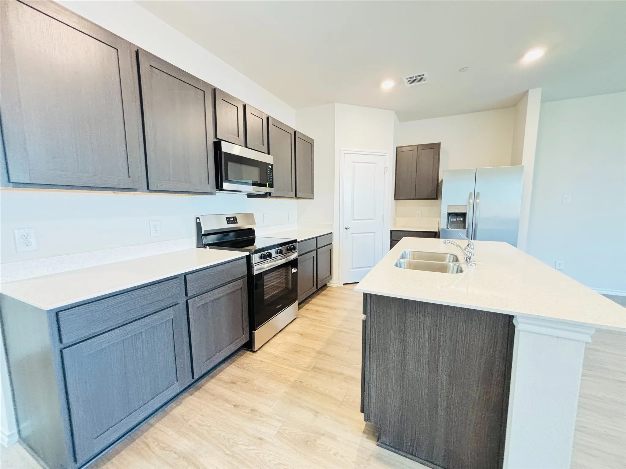 Modern kitchen featuring wood-finish flooring, white countertops, dark wood-finish cabinetry, stainless steel appliances, and a central island with a double basin sink.